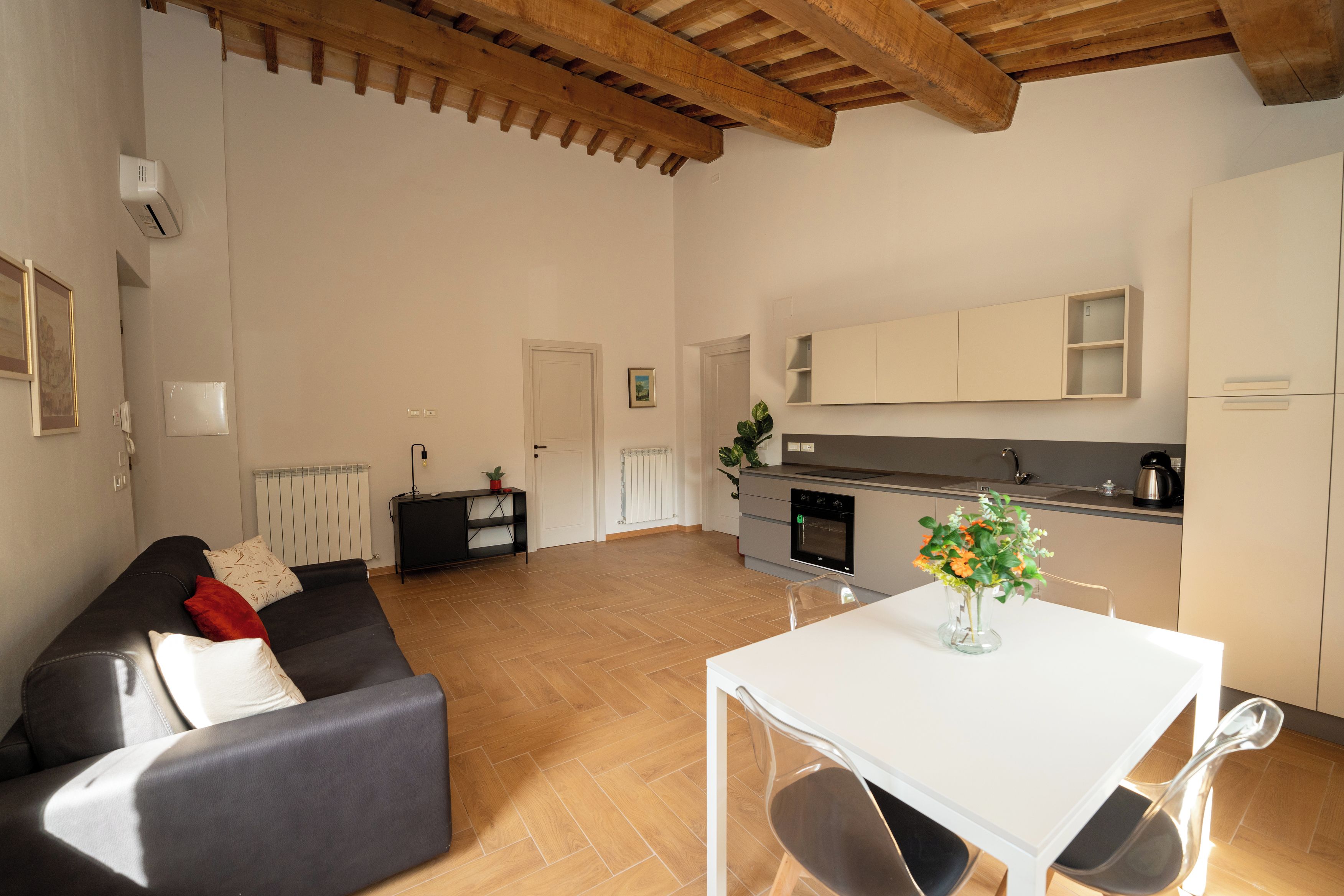 A modern living room with a sofa, a dining table with chairs, a kitchen area with a stove and cabinets, and a radiator.