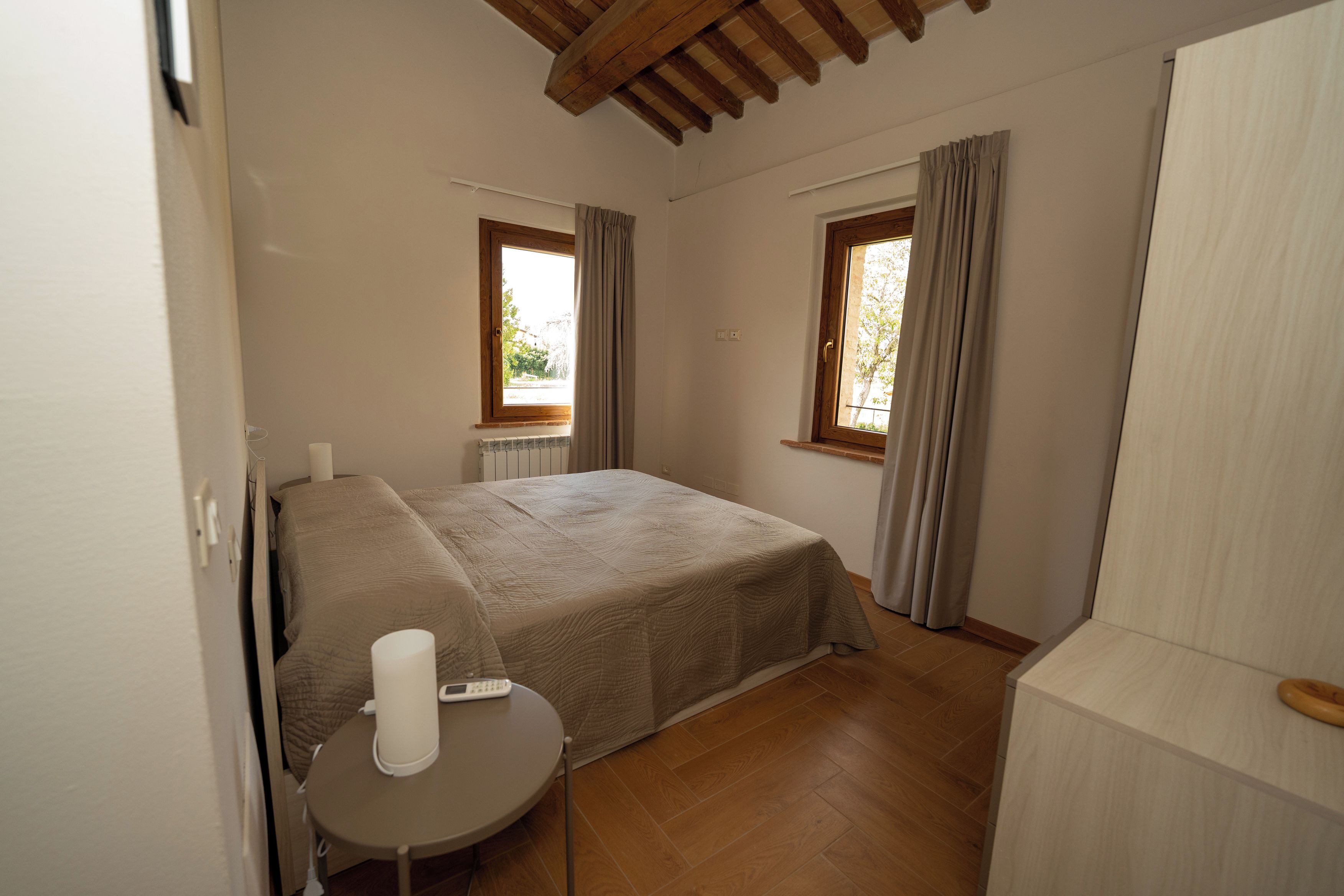 Quarto com cama, mesa de cabeceira e duas janelas com cortinas.