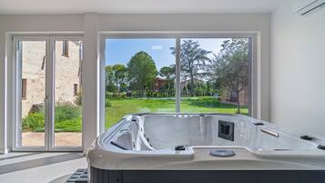 Un baño moderno con jacuzzi, ventanas amplias y vistas a un jardín y edificios.
