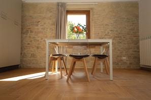 Un área de comedor con una mesa y sillas, una ventana con cortinas, y vista a follaje exterior.
