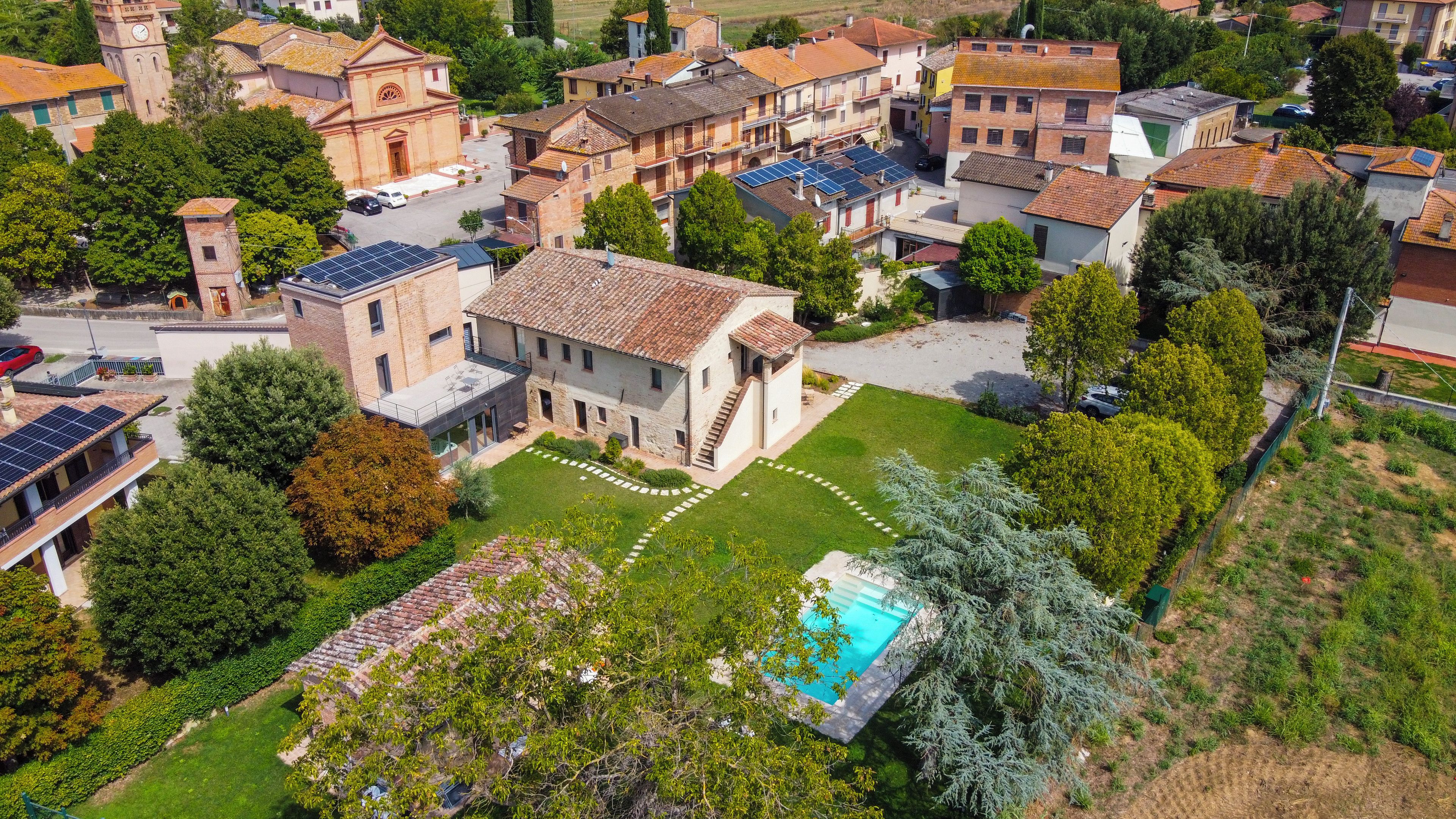 An aerial view of a residential area with a prominent building featuring a bell tower, solar panels, and a swimming pool.