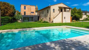 Une piscine dotée d’une terrasse en pierre, entourée d’une pelouse et bordée d’un bâtiment à la façade en pierre et au clocher.