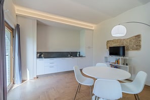 A modern kitchen with white cabinetry, a dining area with a round table and white chairs, a television on a shelf, and a stone wall feature.
