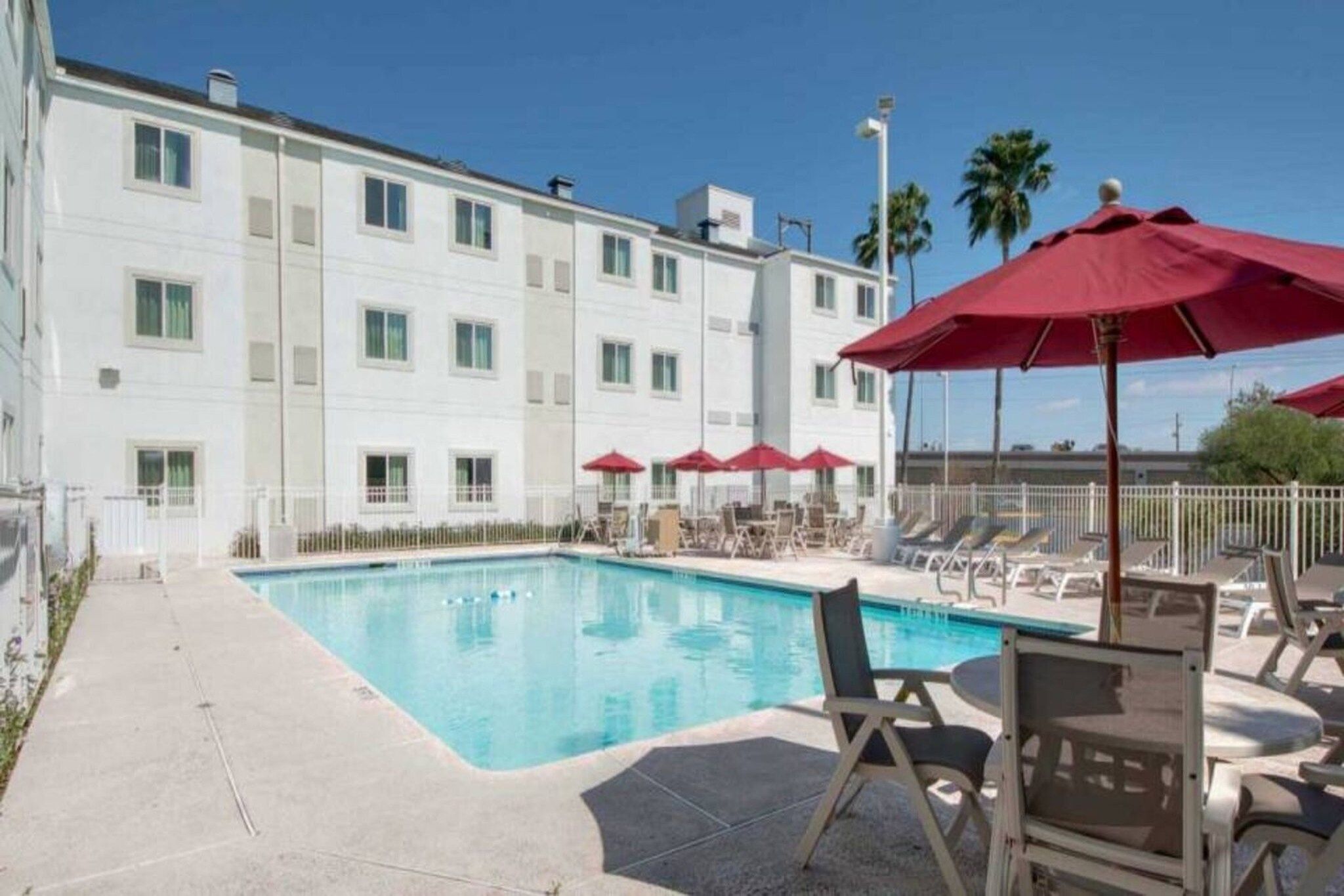 A hotel pool area with a rectangular pool, lounge chairs, and red umbrellas.
