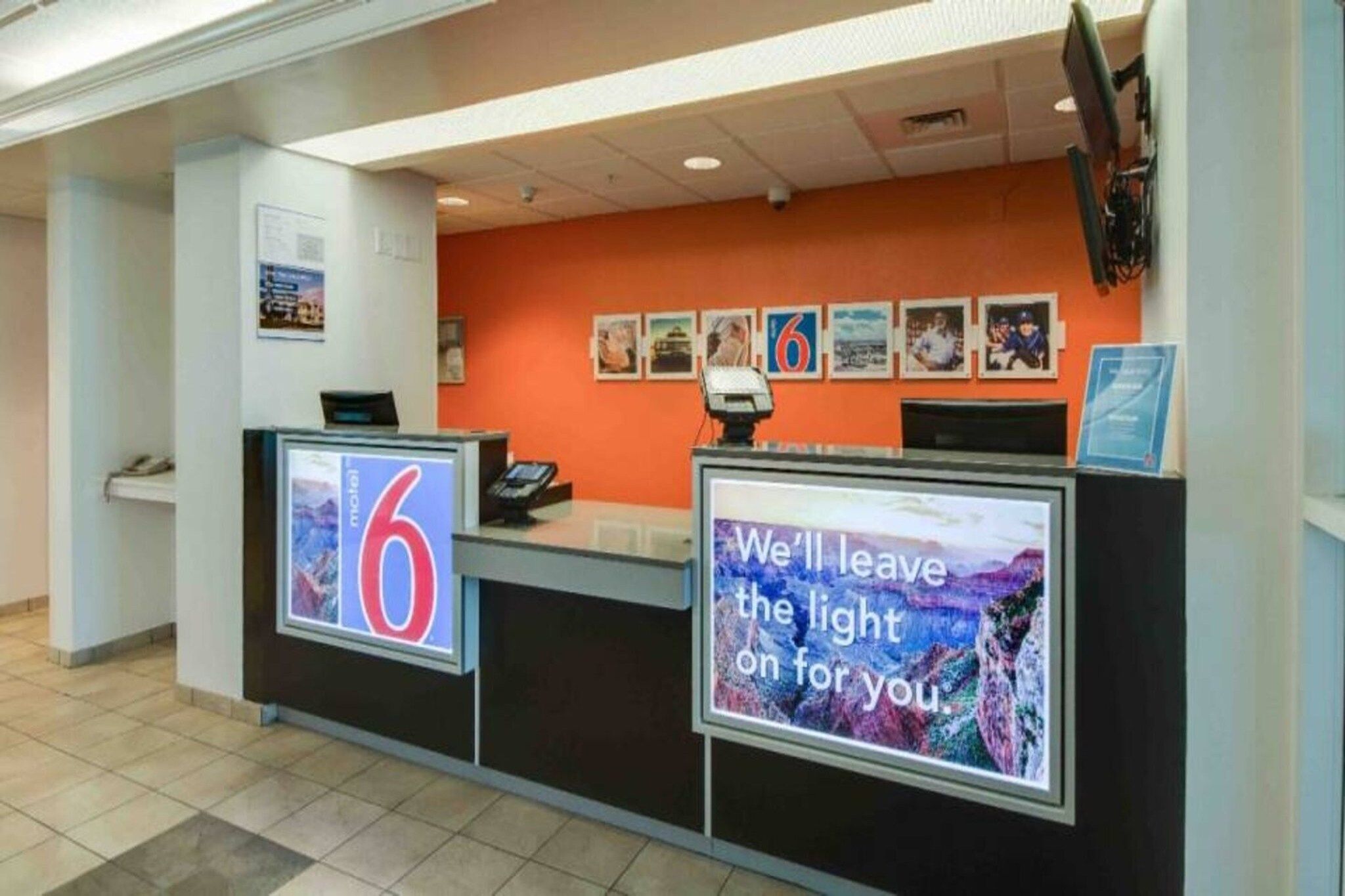 A hotel reception desk with a large number '6' sign, a digital display with a scenic message, and a series of framed pictures on the wall.