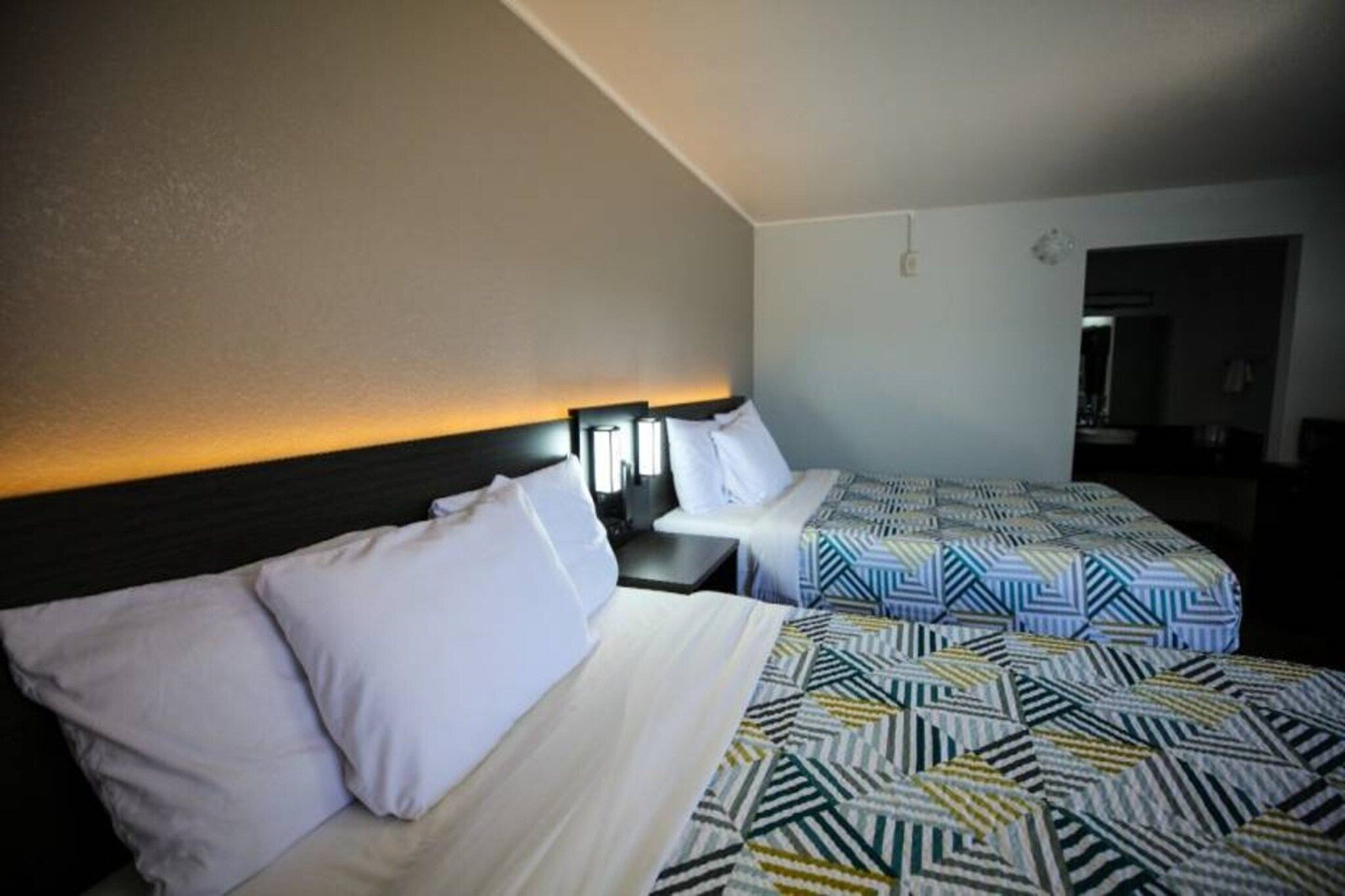 Una habitación de hotel con dos camas, cada una con cabecero y almohadas. Hay una mesita de noche entre las camas con una lámpara encima.