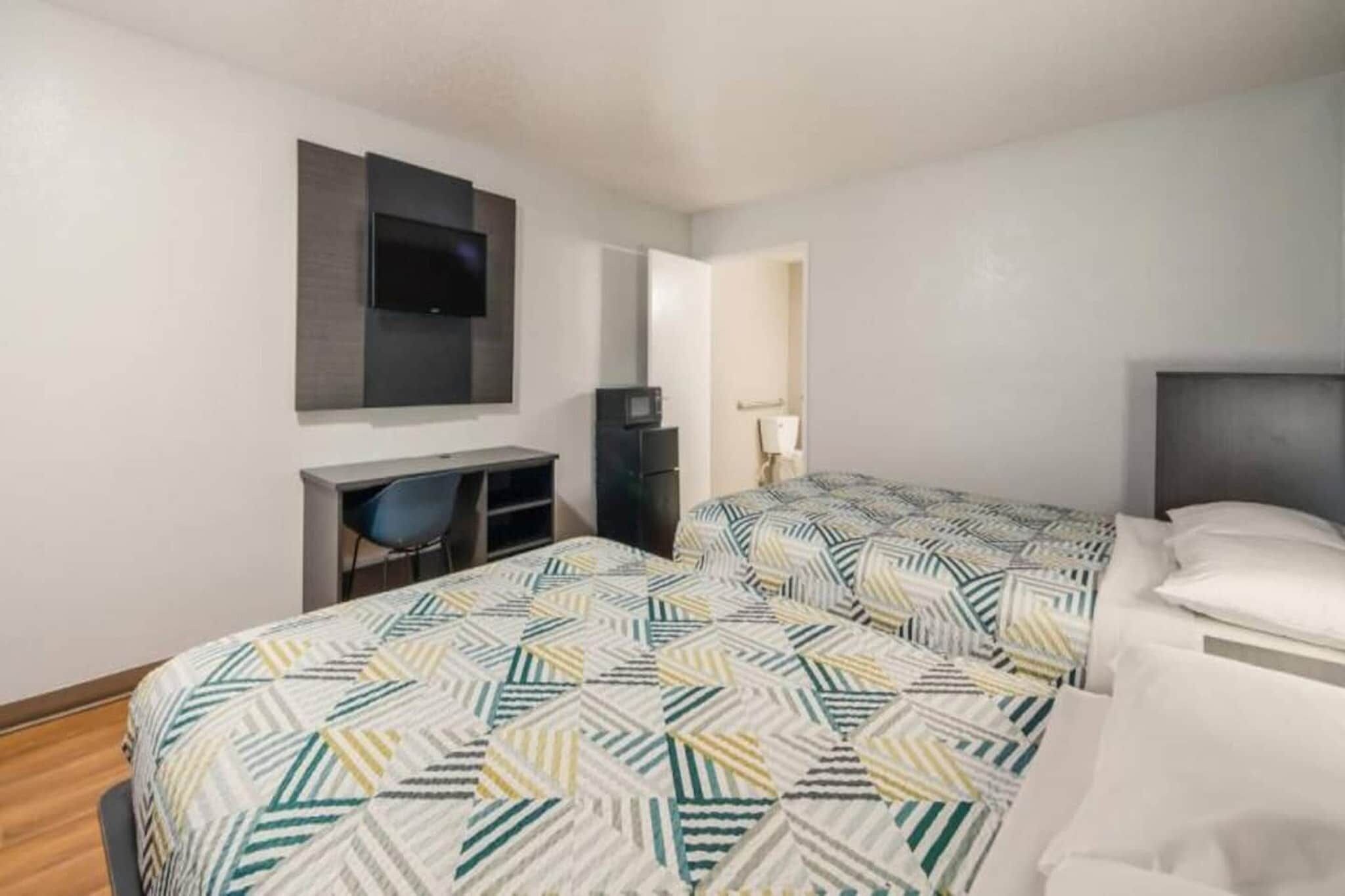 A hotel room with two beds, a desk, a chair, a TV, and a lamp.