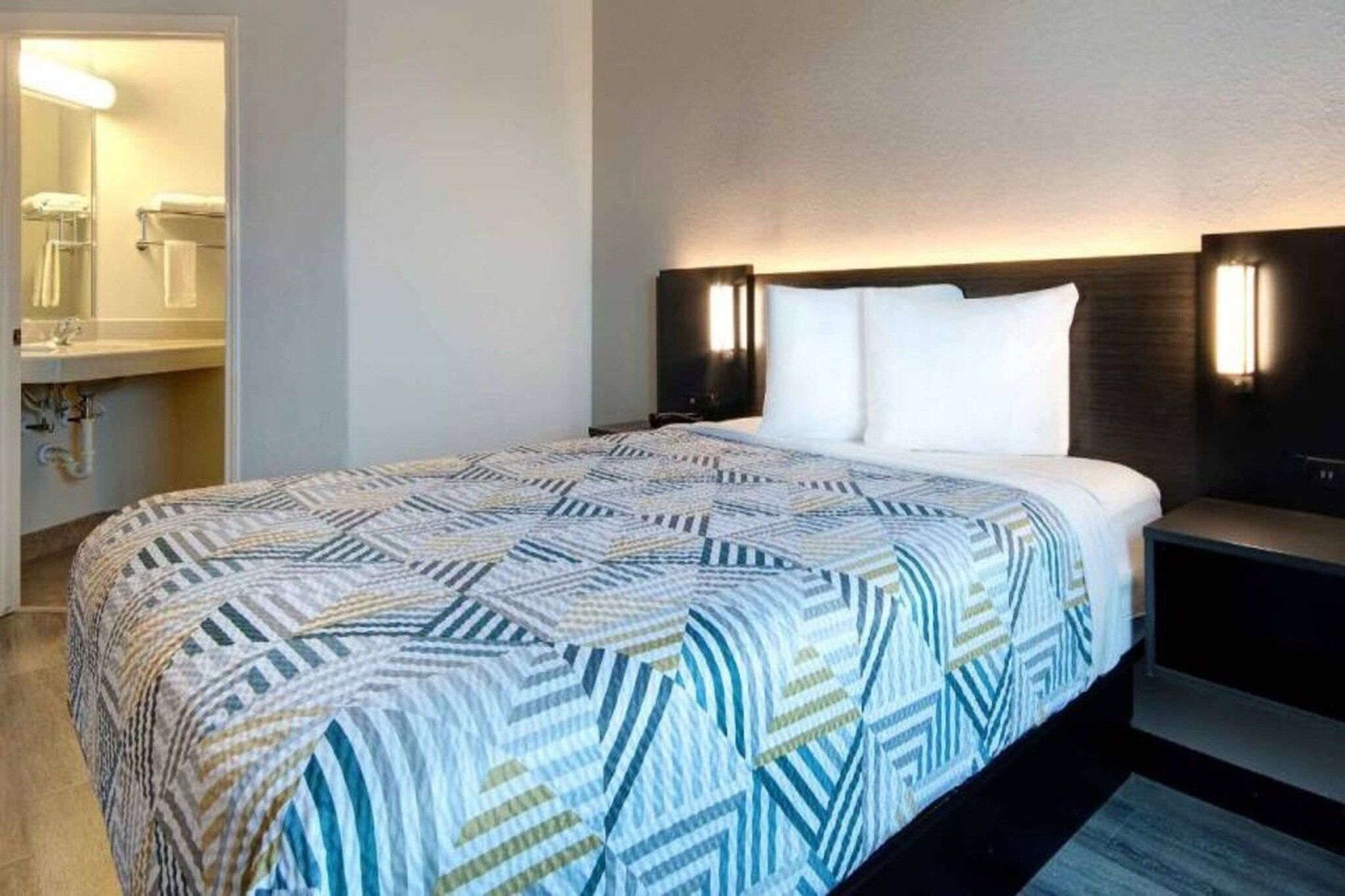 A hotel room with a large bed, a nightstand, a bedside table, a wall-mounted lamp, and a bathroom visible through an open door.