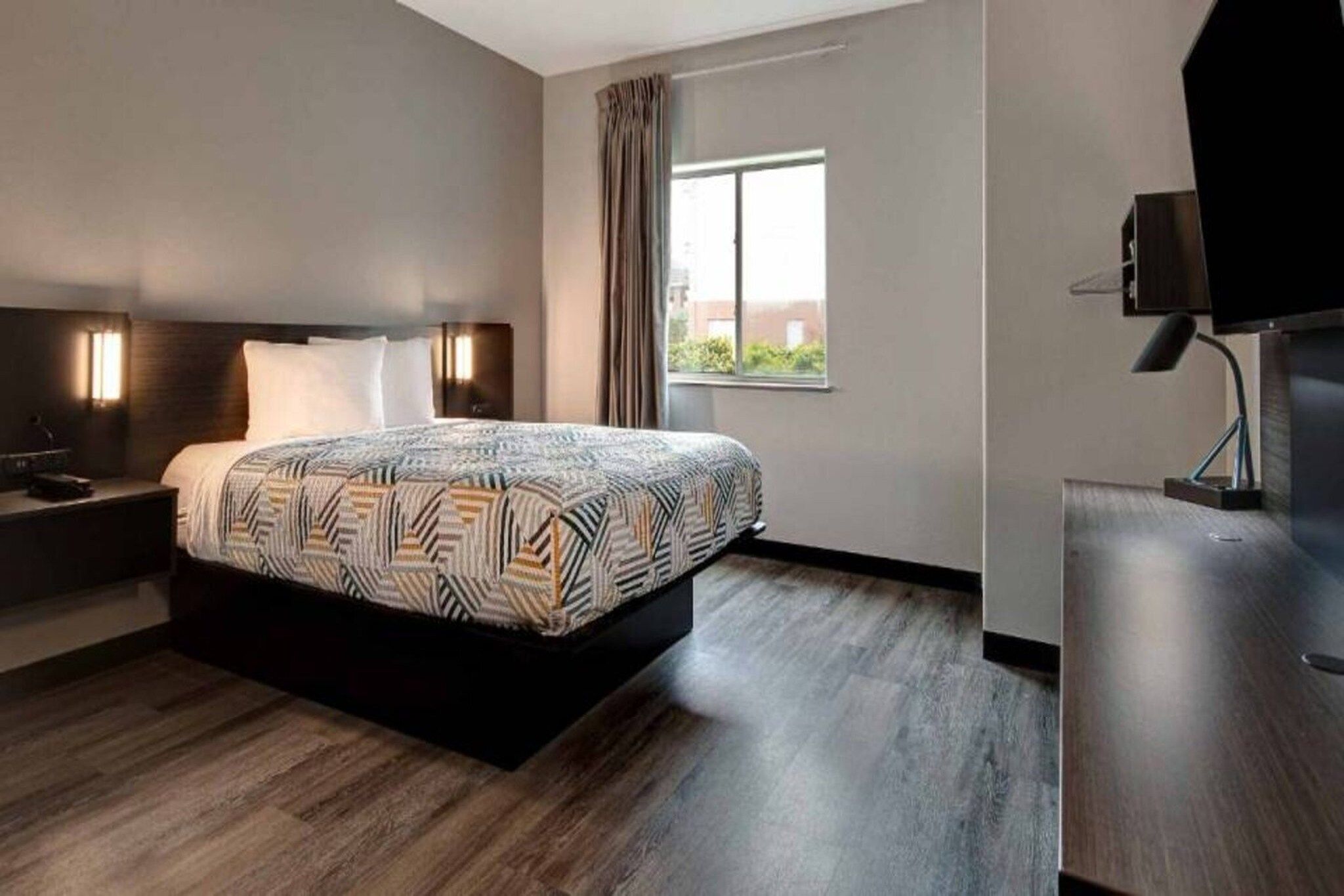 Ein modernes Hotelzimmer mit einem großen Bett, einem Nachttisch, einem Schreibtisch mit Stuhl, einem Fernseher und einem Fenster mit Vorhängen.
