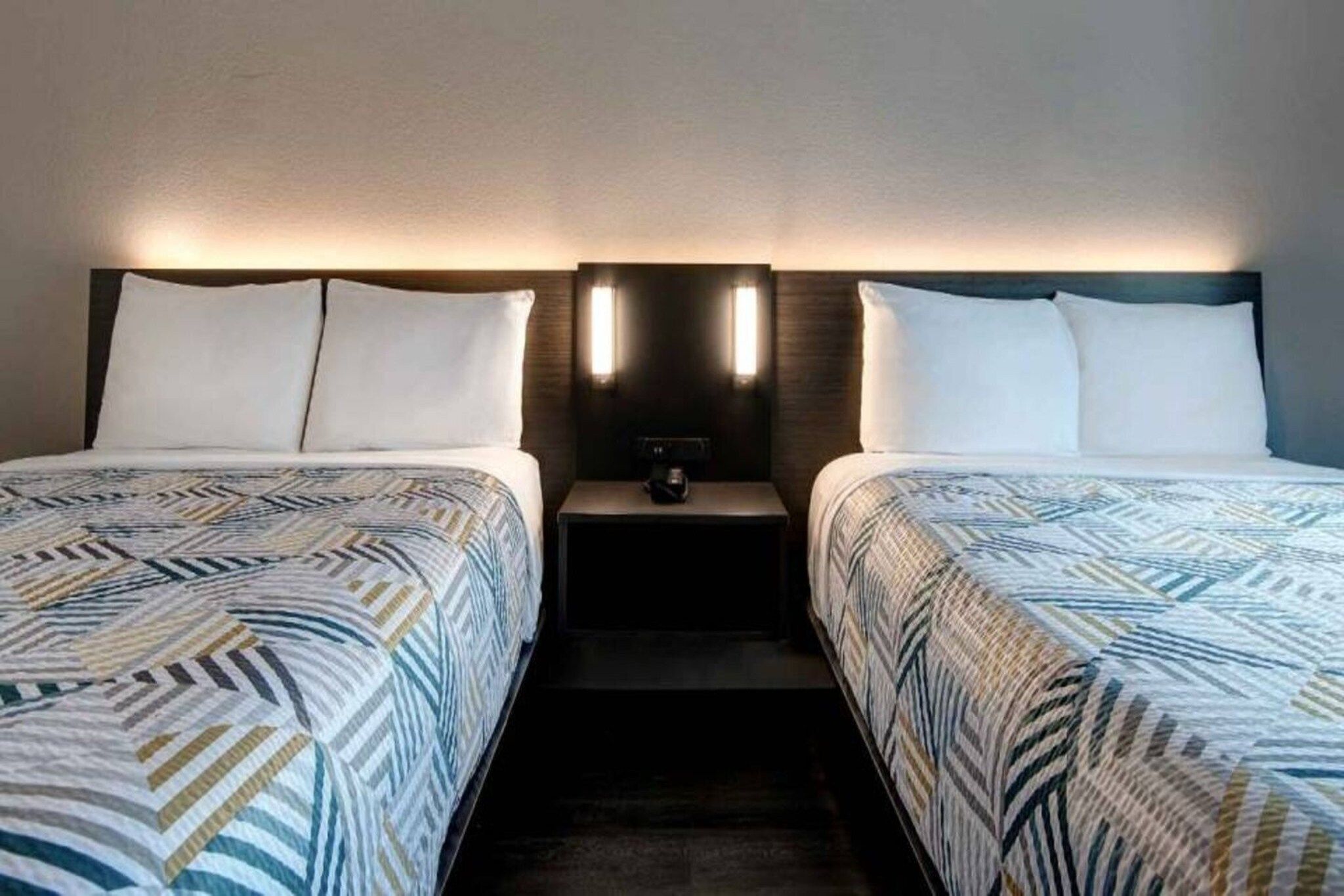 A hotel room with two beds, each with patterned bedding, a nightstand with a lamp, and wall-mounted lights.