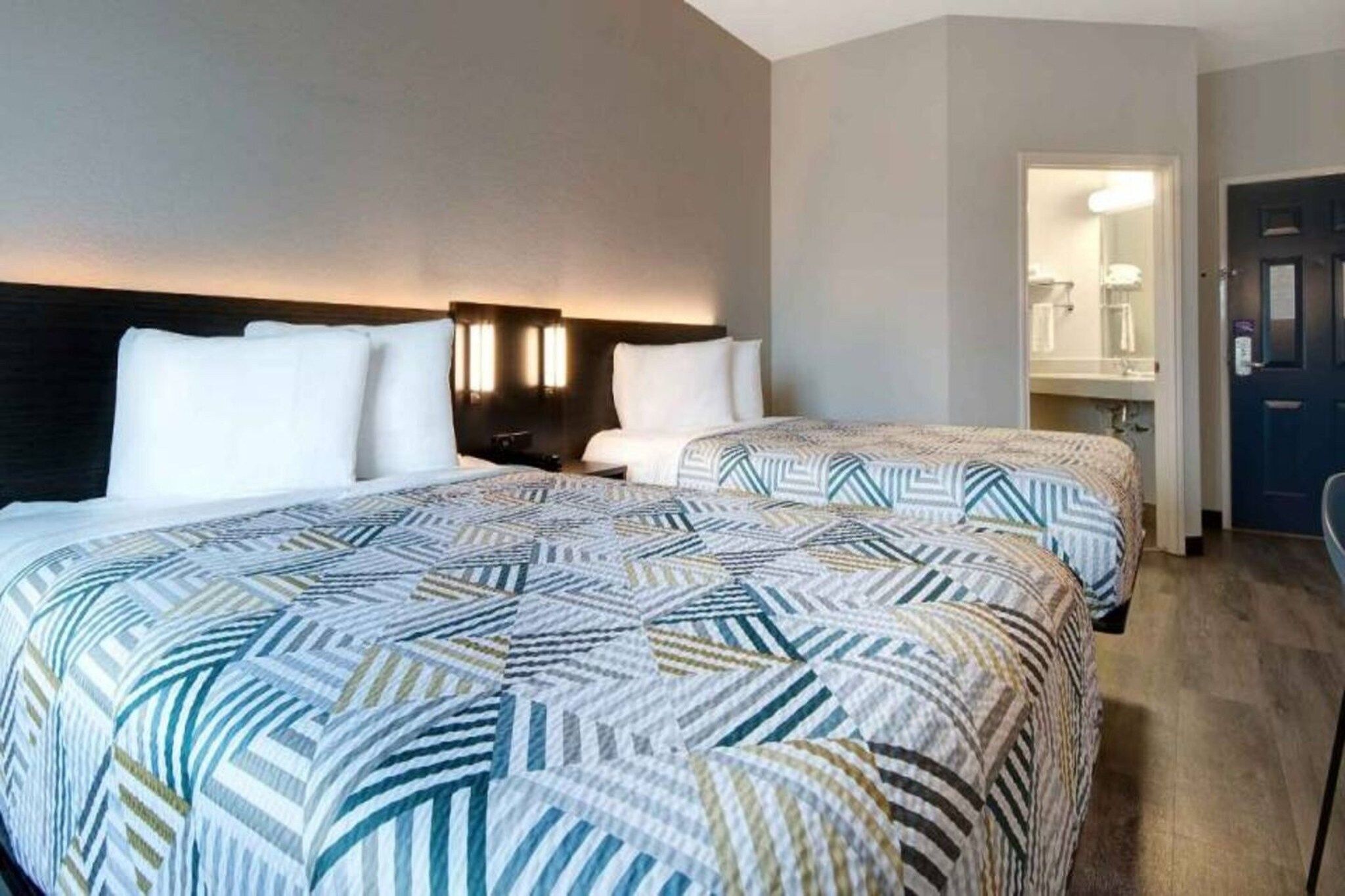 A hotel room with two beds, a dark headboard, a nightstand, and a bathroom visible through an open door.