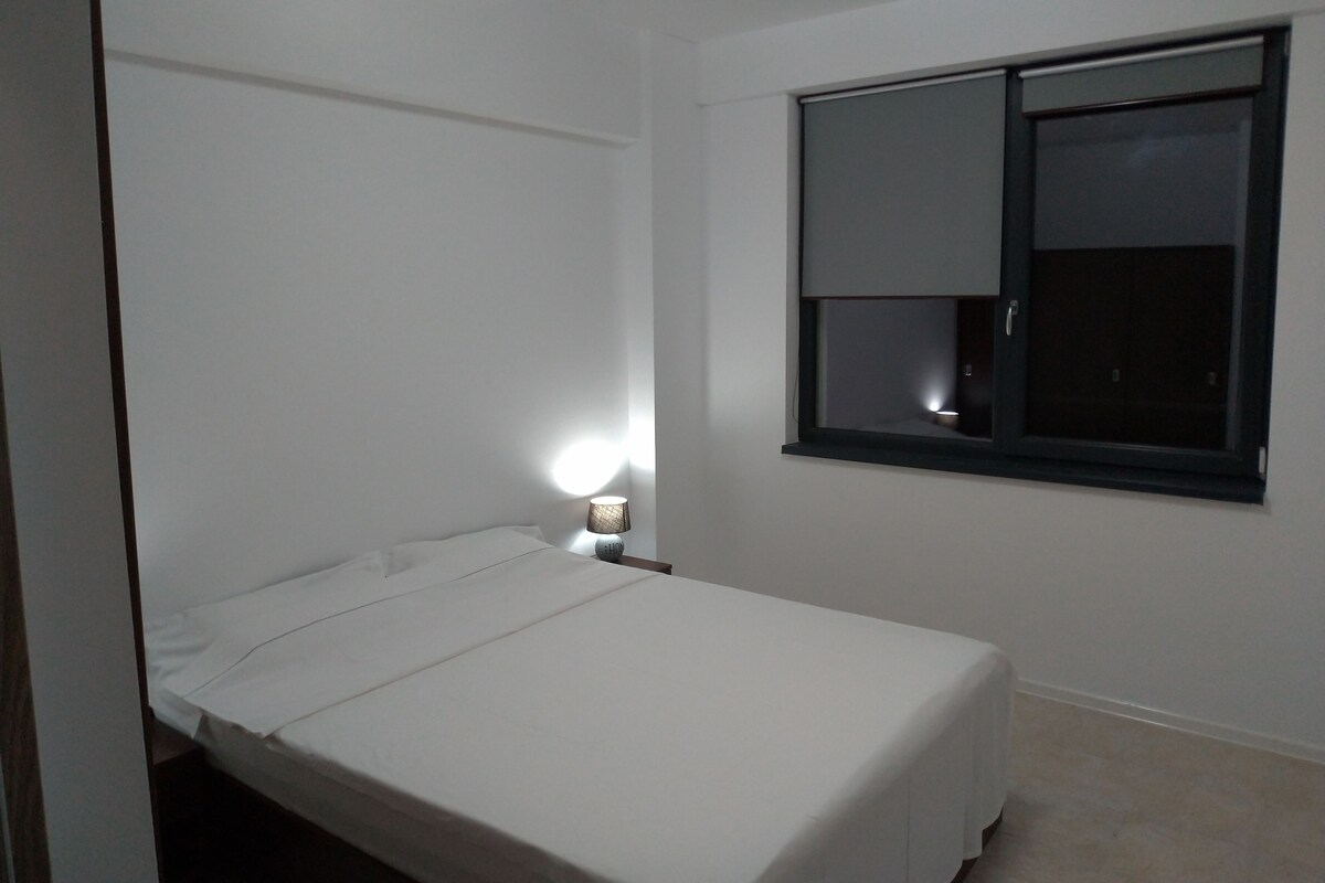 A small, single-bed room with a window, a bedside lamp, and a door.