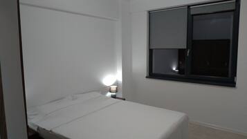 A small, single-bed room with a window, a bedside lamp, and a door.
