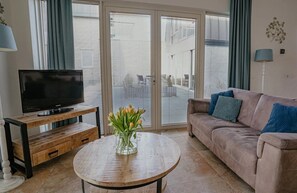 A living room with a television on a wooden stand, a sofa with cushions, a coffee table with a vase of flowers, and a sliding glass door leading to an outdoor area. - Dune apartment Flat Fish in Egmond aan Zee for 3 guests, with a sun terrace, sea views and direct beach access. (Egmond aan Zee)