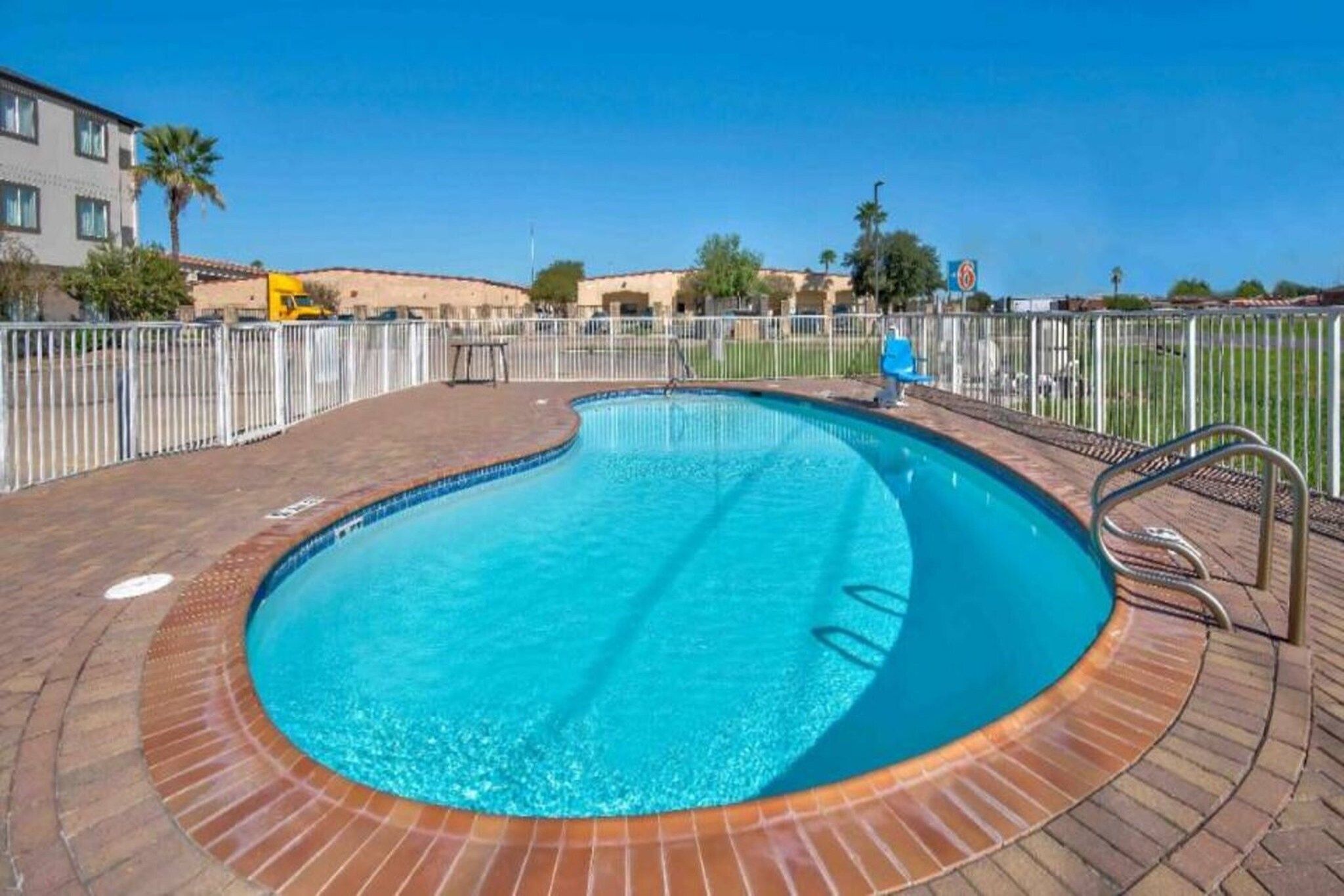 A swimming pool with a brick deck, surrounded by a white fence, and a clear blue sky in the background.