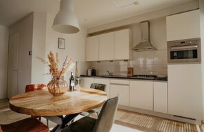 A modern kitchen with white cabinets, a round wooden dining table, and a hanging light fixture. - Sea-view apartment Sea Bass in Egmond aan Zee for 4 guests, with 2 bedrooms and two sunny terraces. (Egmond aan Zee)