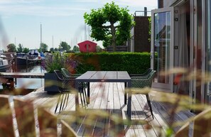 A wooden deck with a table and chairs, a red birdhouse, and a hedge. - Waterlodge in Uitgeest for 4 guests with a spacious waterside terrace, a fully equipped kitchen, two bedrooms and a private parking space. (Uitgeest)