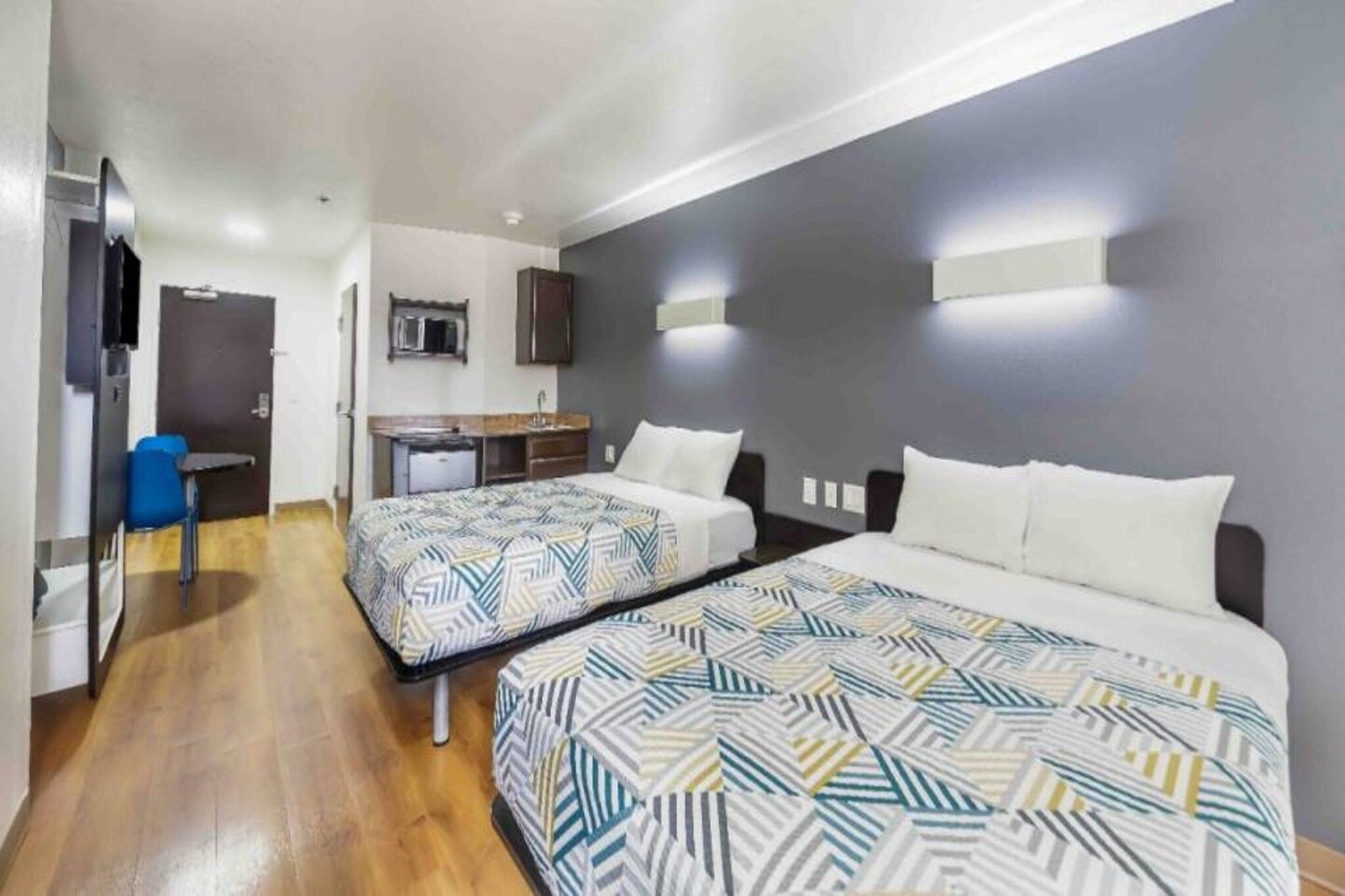 Habitación de hotel con dos camas, un escritorio, una silla y una kitchenette pequeña.
