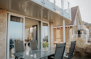 A covered outdoor dining area with a table and chairs, a glass door leading inside, and a view of a brick building. - Beach apartment Lobster in Egmond aan Zee for 6 guests, with 3 bedrooms, a terrace and views of the sea and boulevard. (Egmond aan Zee)