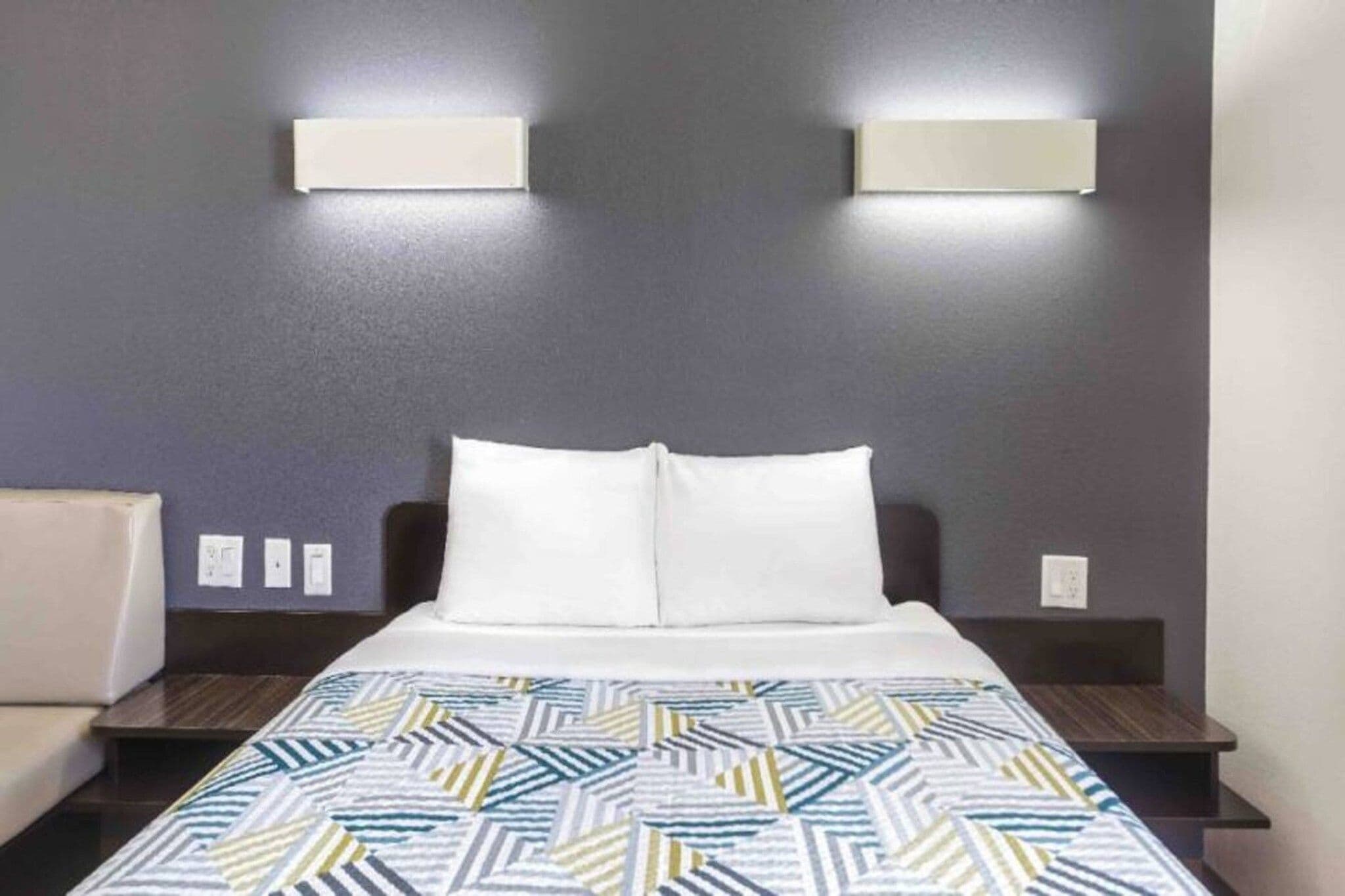 Habitación de hotel con una cama, dos mesitas de noche, un buró, una colcha estampada y dos lámparas fijadas a la pared.