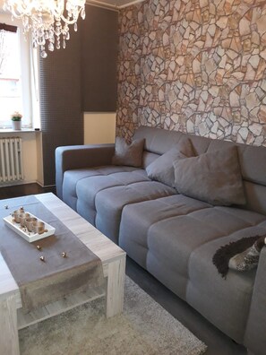 A living room with a grey sofa, a white coffee table with decorative items, a stone wall, and a chandelier. - Eifelglück House (Großlittgen)