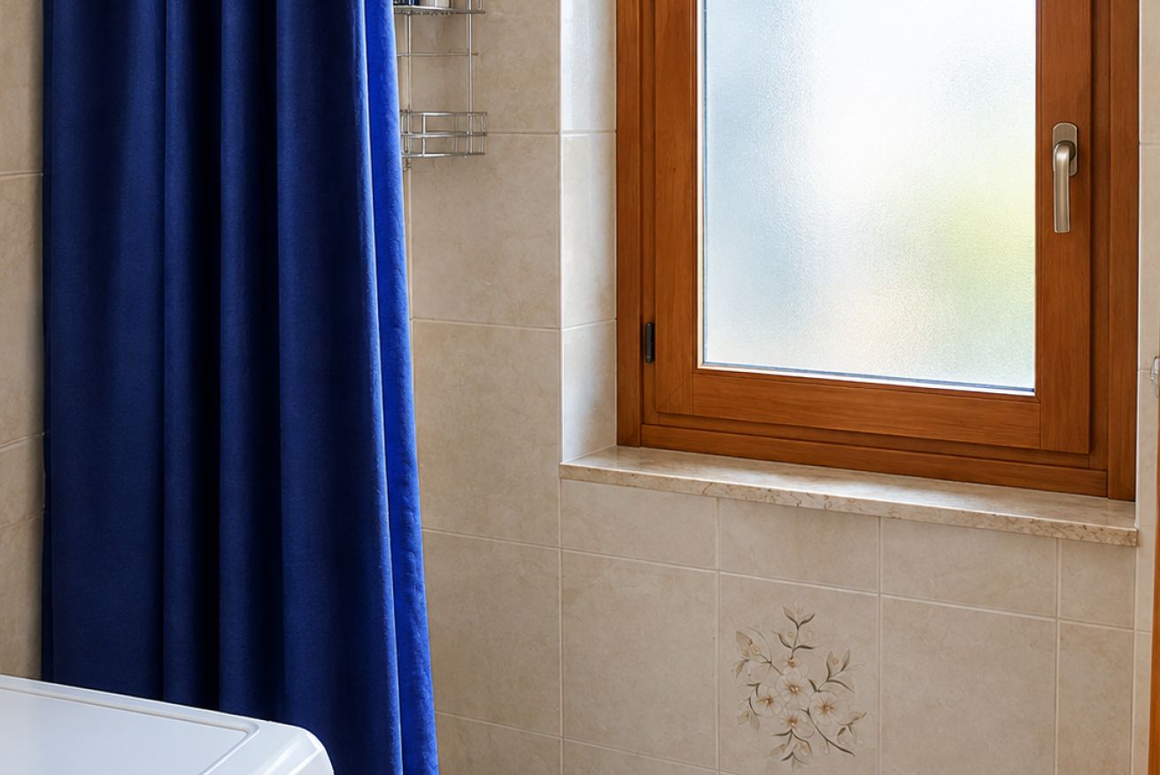Un baño con cortina de ducha azul, ventana con marco de madera y pared de azulejos con diseño floral.