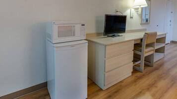 A compact room with a kitchenette area featuring a microwave and a small refrigerator. There is a desk with a chair and a television on top.