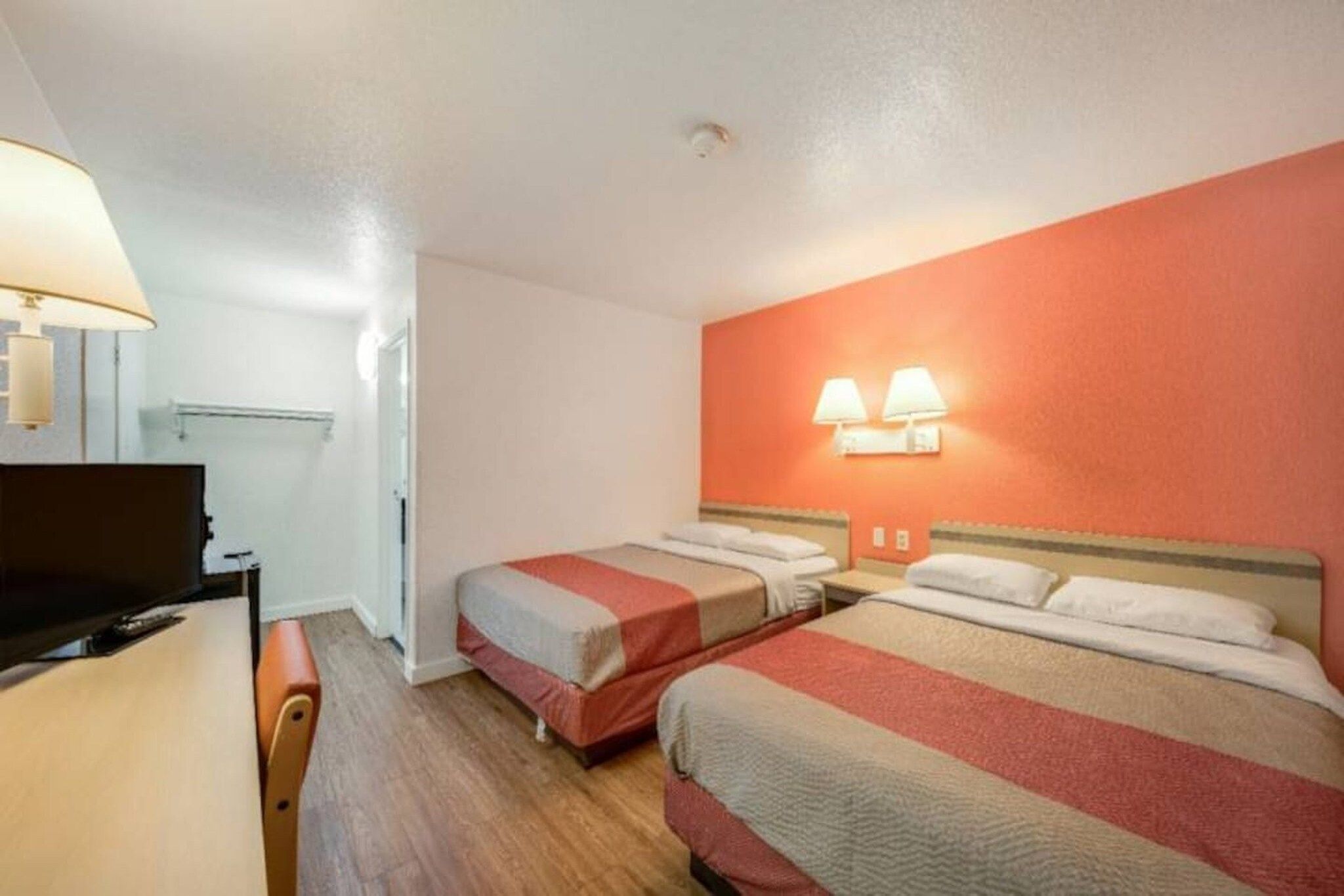 Quarto de hotel com duas camas, uma escrivaninha, uma televisão e um candeeiro fixado na parede.
