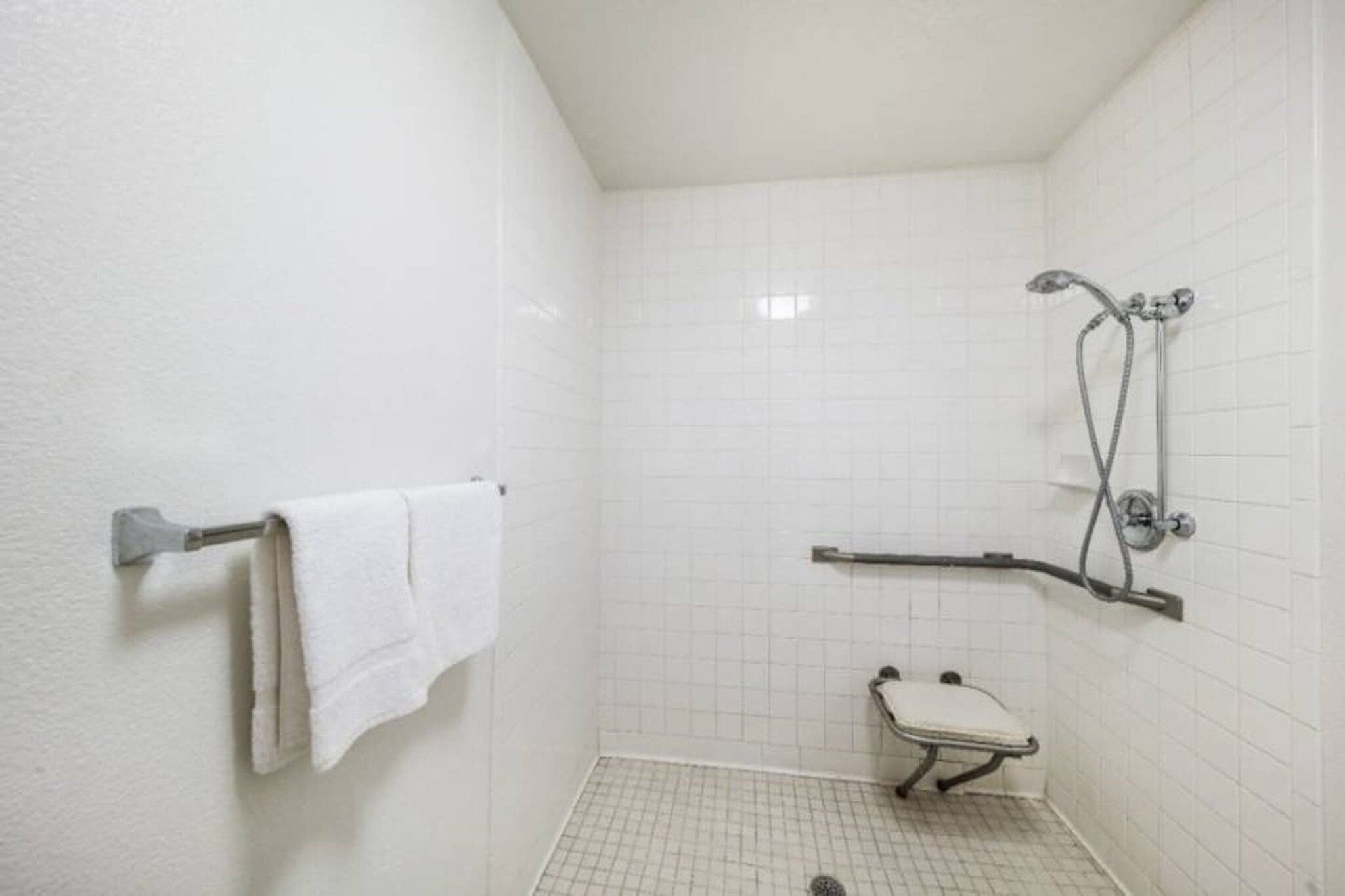 Une petite salle de douche aux murs recouverts de carreaux blancs, équipée d’une barre d’appui et d’une serviette blanche posée sur un porte-serviettes.