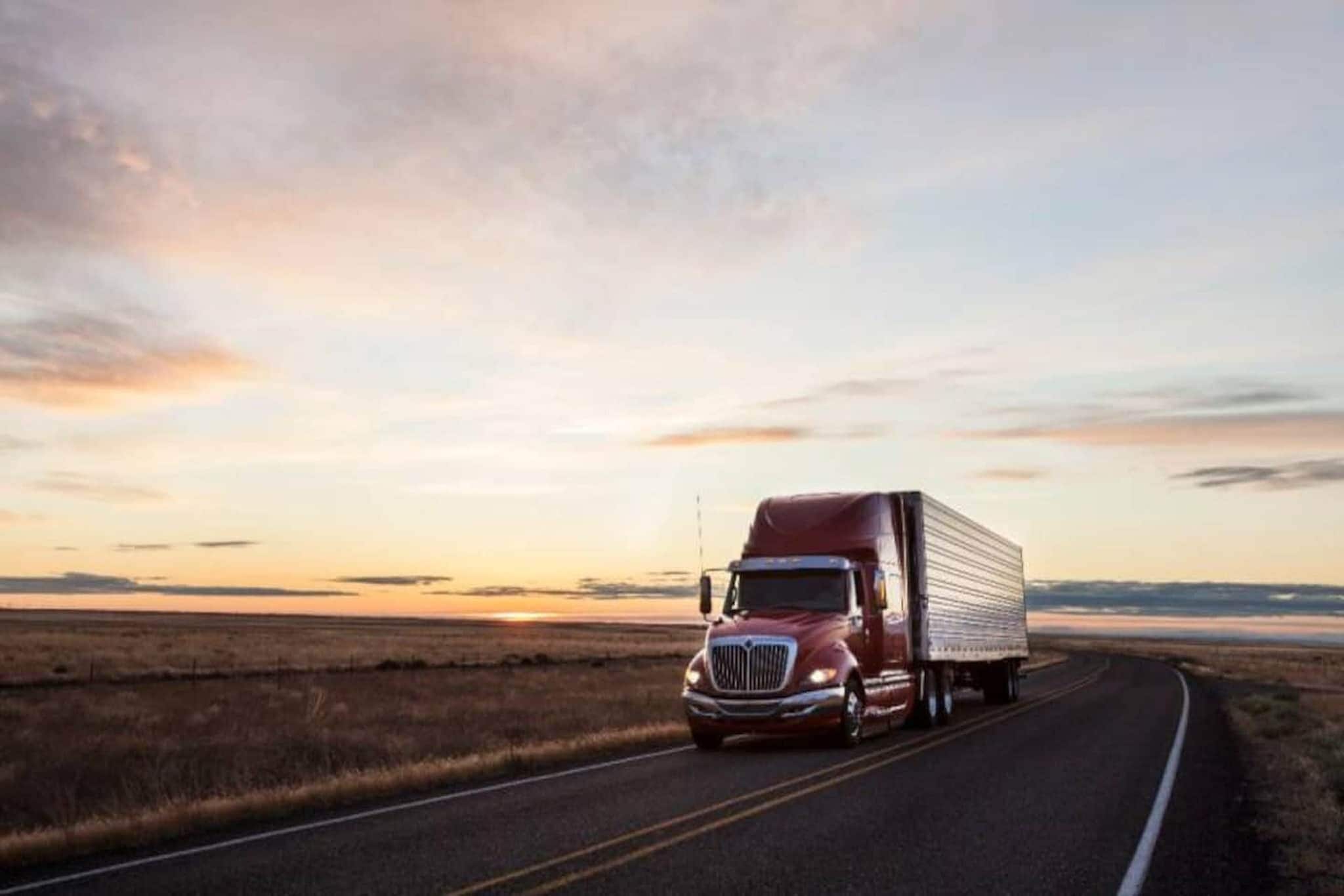 Un camion semi-remorque rouge sur une route à deux voies, avec un coucher de soleil en arrière-plan.