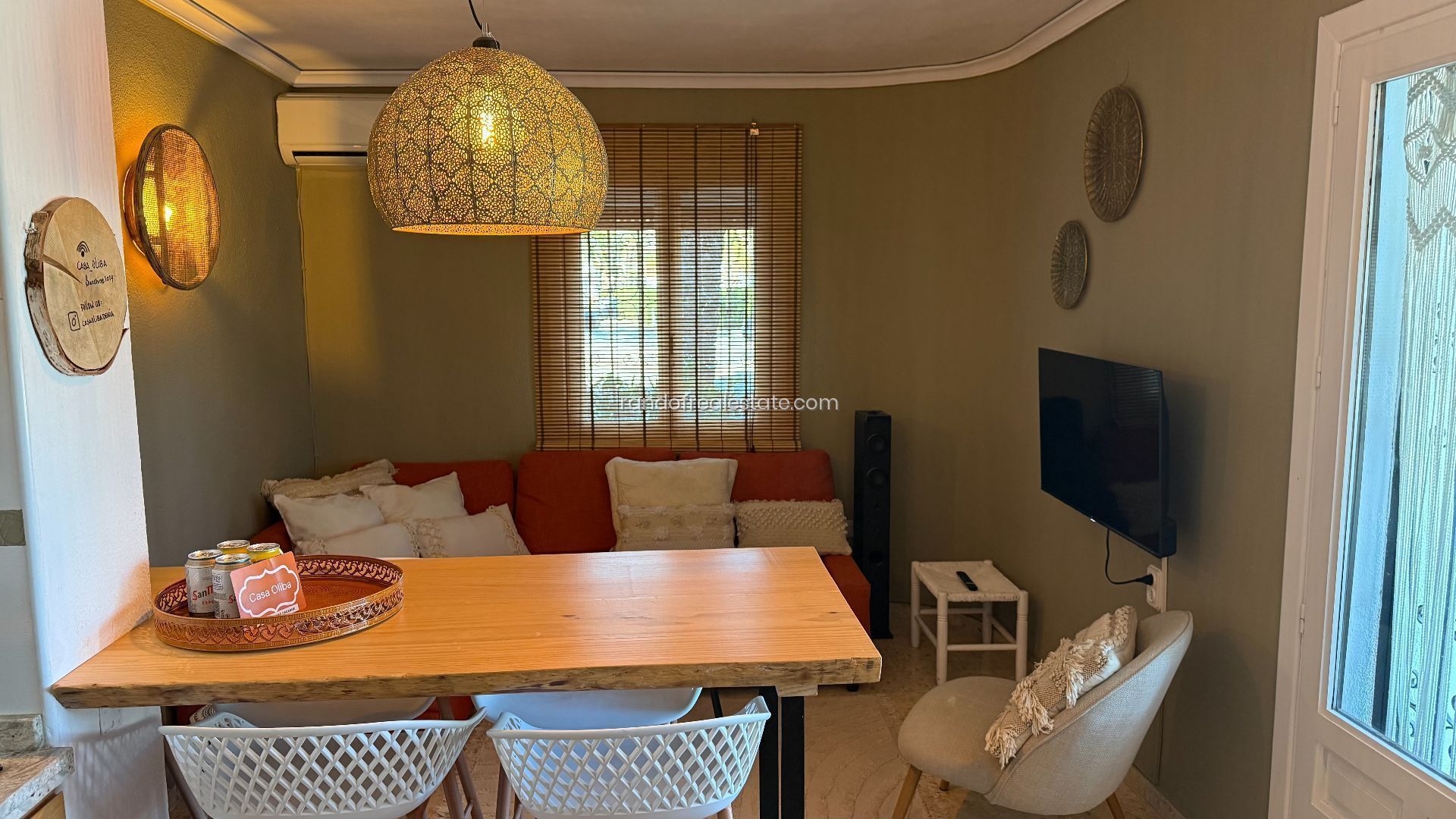 Una sala acogedora con una mesa de madera, sillas blancas, un sofá con cojines rojos, un televisor y una lámpara colgante decorativa.