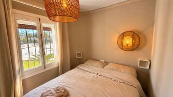 A bedroom with a bed, a window with curtains, a wall-mounted light fixture, and a small bedside table.
