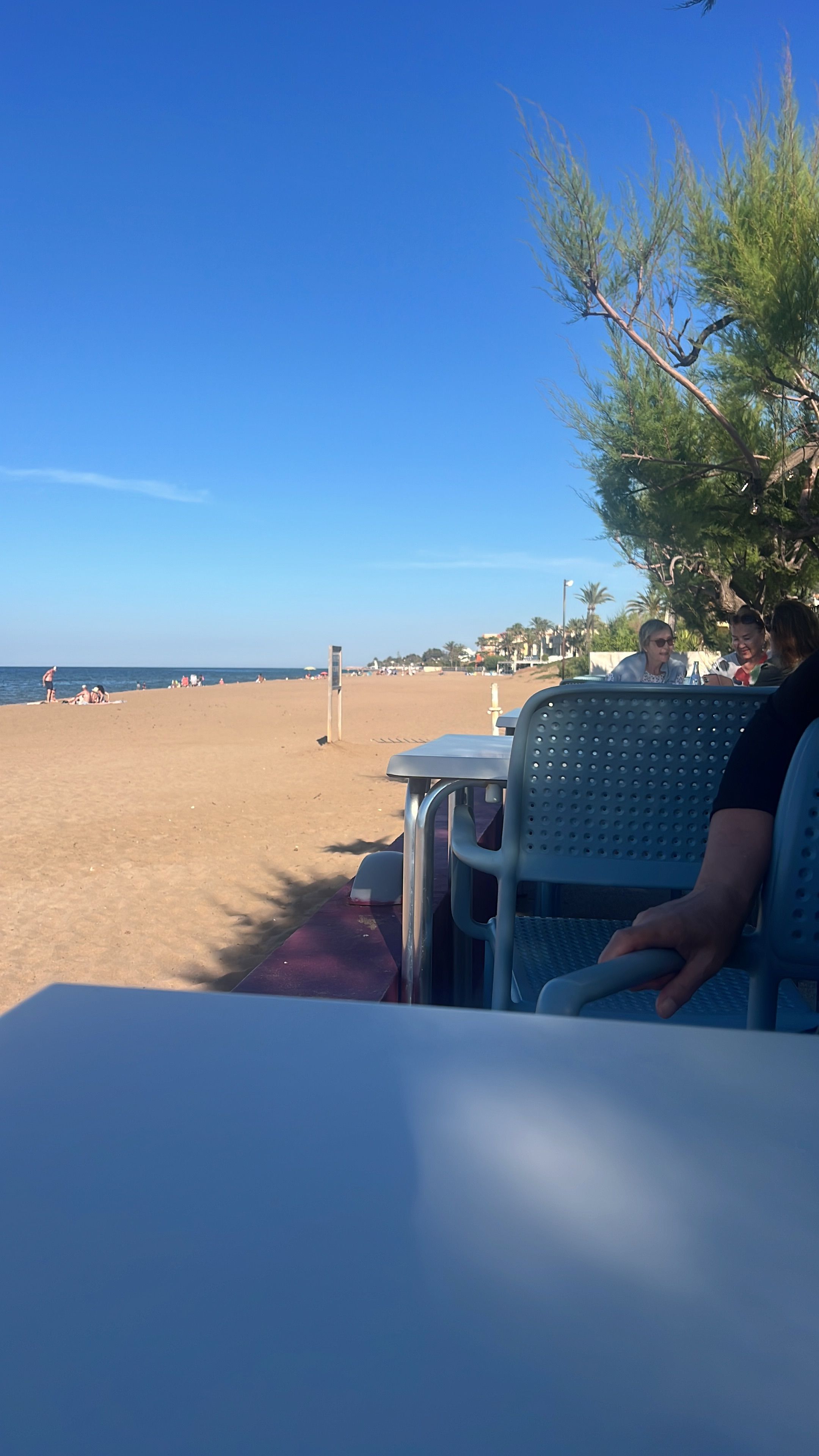 Una mesa con sillas frente a la playa, un cielo azul despejado y arena al fondo.