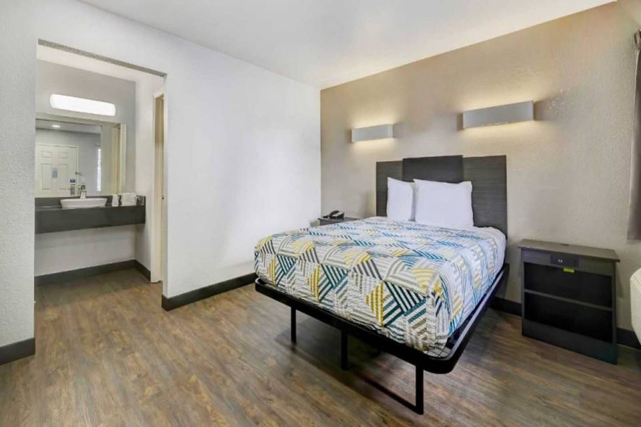 Una habitación de hotel con una cama, una mesita de noche y un baño visible a través de una puerta abierta.