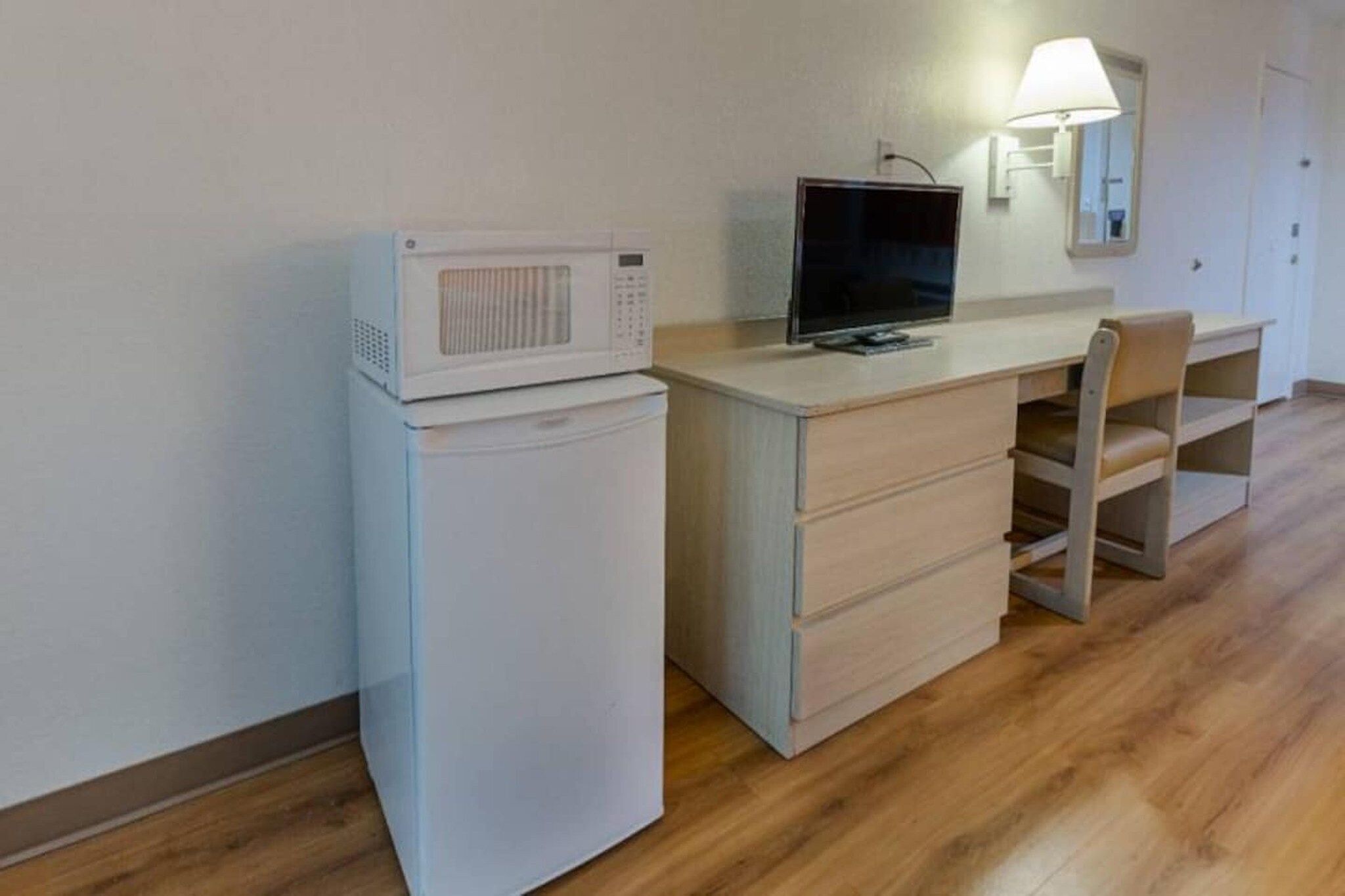 A compact room with a kitchenette area featuring a microwave and a small refrigerator. There is a desk with a chair and a television on top.