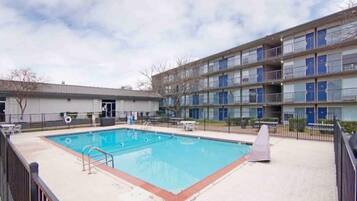 A swimming pool with a metal fence, a covered seating area, and a multi-story building with balconies in the background.