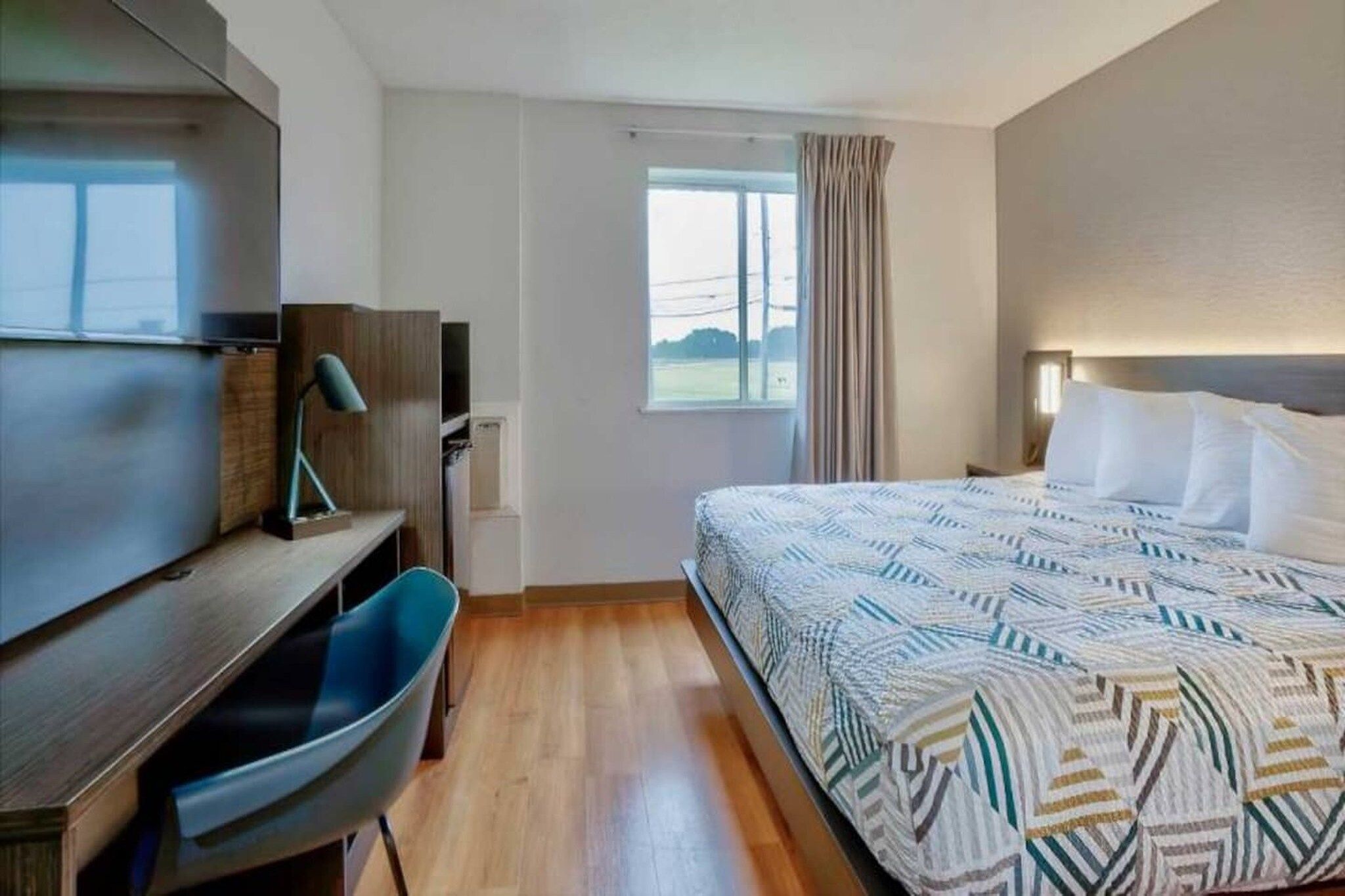 Ein Hotelzimmer mit Bett, Schreibtisch, Stuhl, einem Fenster mit Vorhängen und Blick ins Freie.