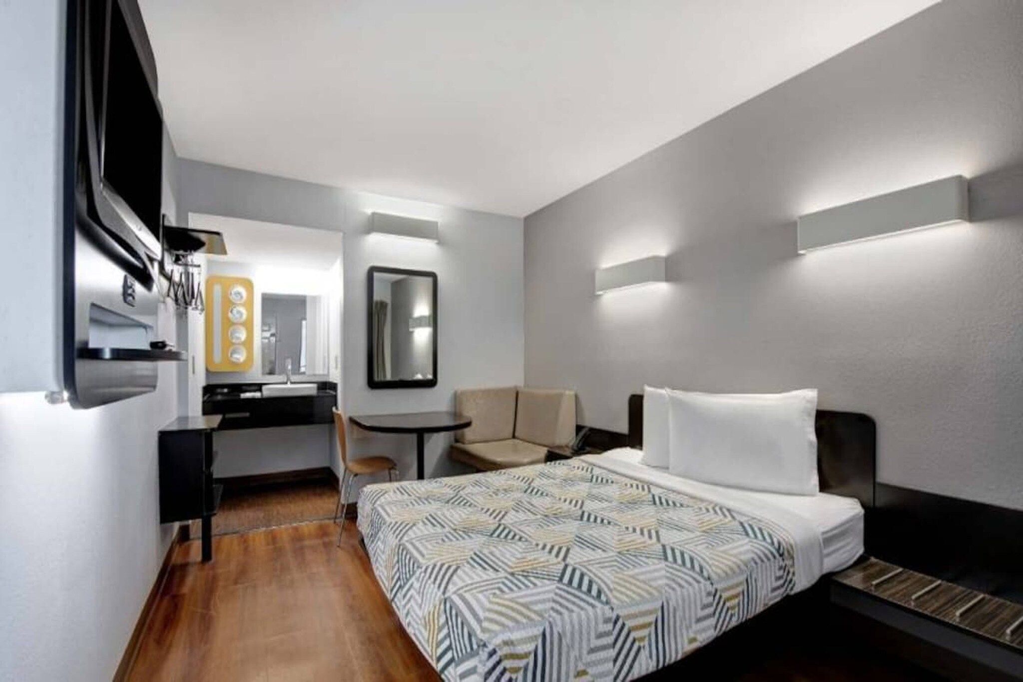 A hotel room with a bed, a desk, a chair, a mirror, and a wall-mounted light fixture.