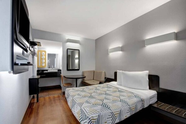A hotel room with a bed, a desk, a chair, a mirror, and a wall-mounted light fixture.