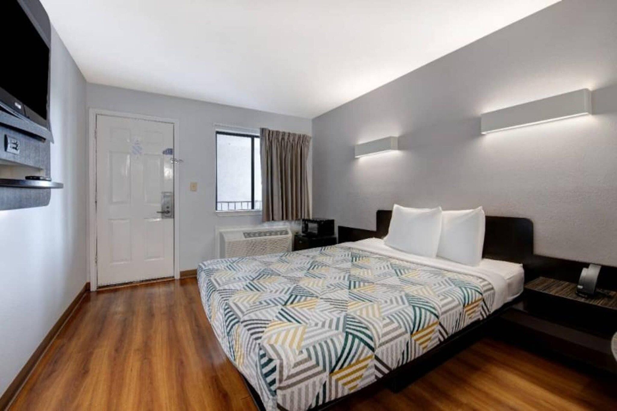 A hotel room with a bed, a desk, a chair, a window with curtains, and a door.