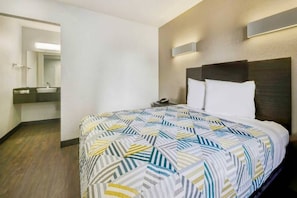 A hotel room with a bed, a nightstand, a mirror, and a bathroom visible in the background.