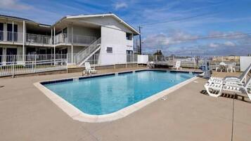 A swimming pool with a white building in the background, a fenced area, and lounge chairs.
