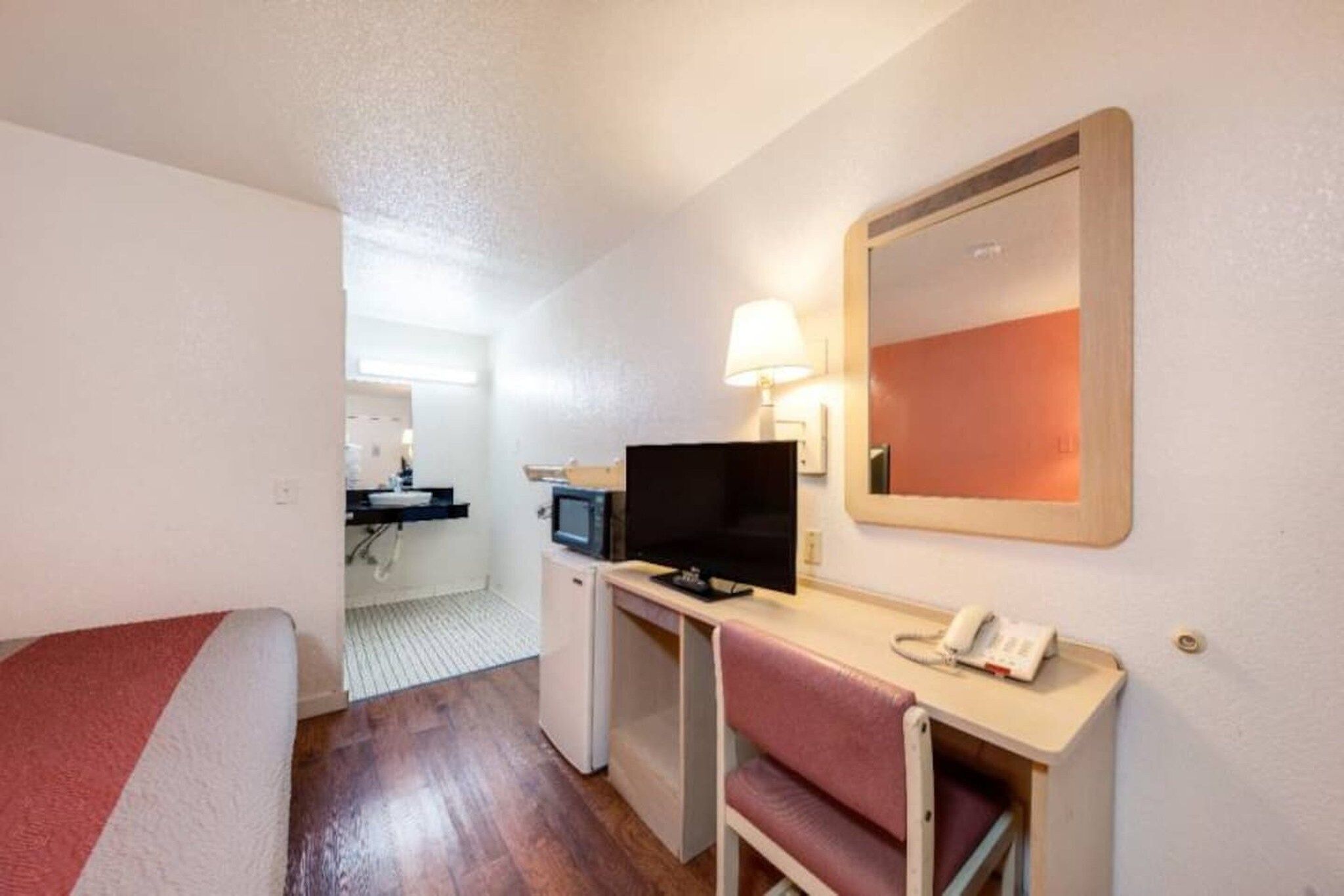 Habitación de hotel con cama, escritorio con computadora y teléfono, kitchenette pequeña y un espejo en la pared.