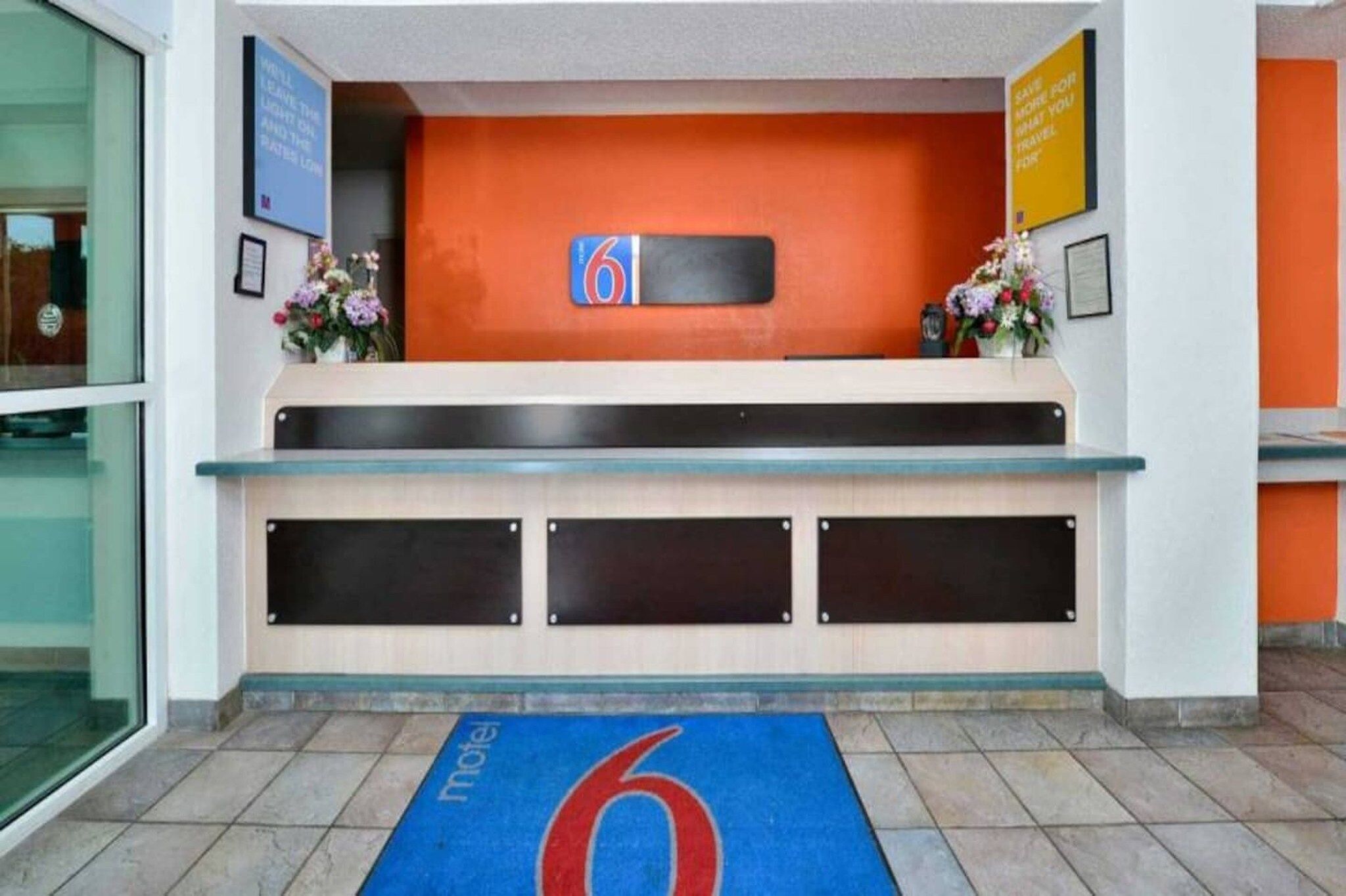 A hotel reception desk with a blue carpet featuring a red number 6, a sign with the number 6, and a flower arrangement on the counter.
