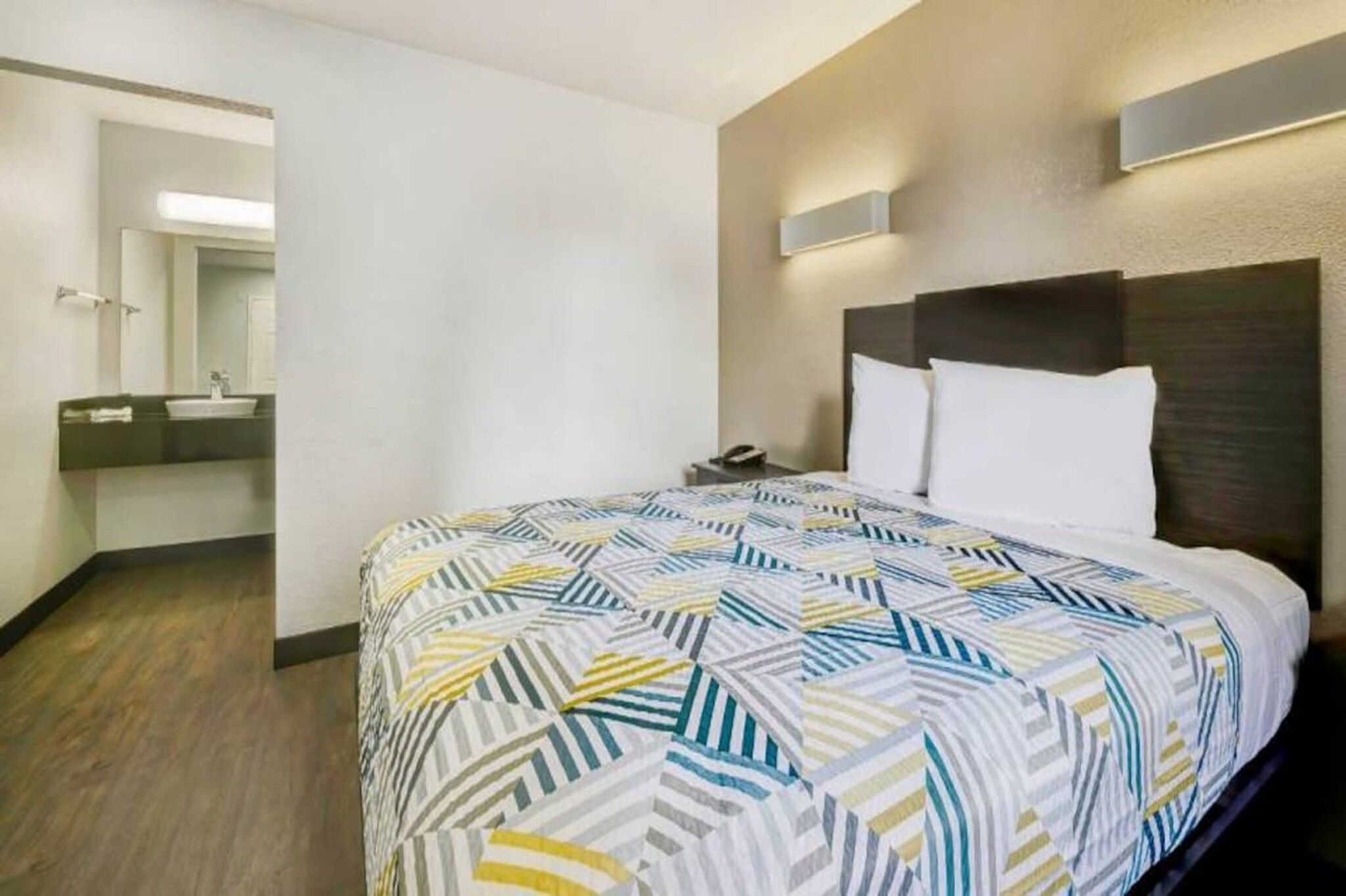 A hotel room with a bed, a nightstand, a mirror, and a bathroom visible in the background.