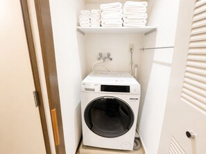 A compact laundry room with a front-loading washing machine, a stacked shelf with folded towels, and a wall-mounted faucet. - Ocean Shinsaibashi Higashi (Osaka)