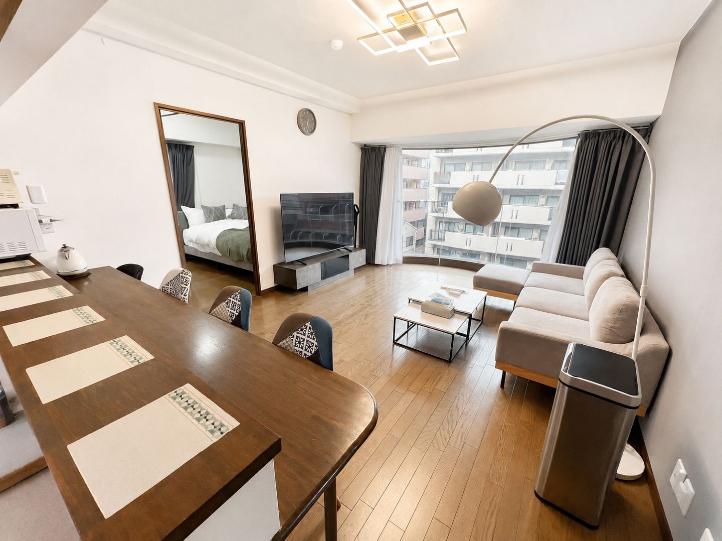 A modern hotel room with a wooden dining table, a sofa, a TV, and a large window offering a city view.