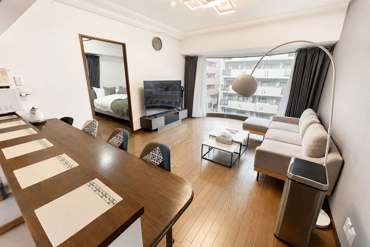 A modern hotel room with a wooden dining table, a sofa, a TV, and a large window offering a city view.