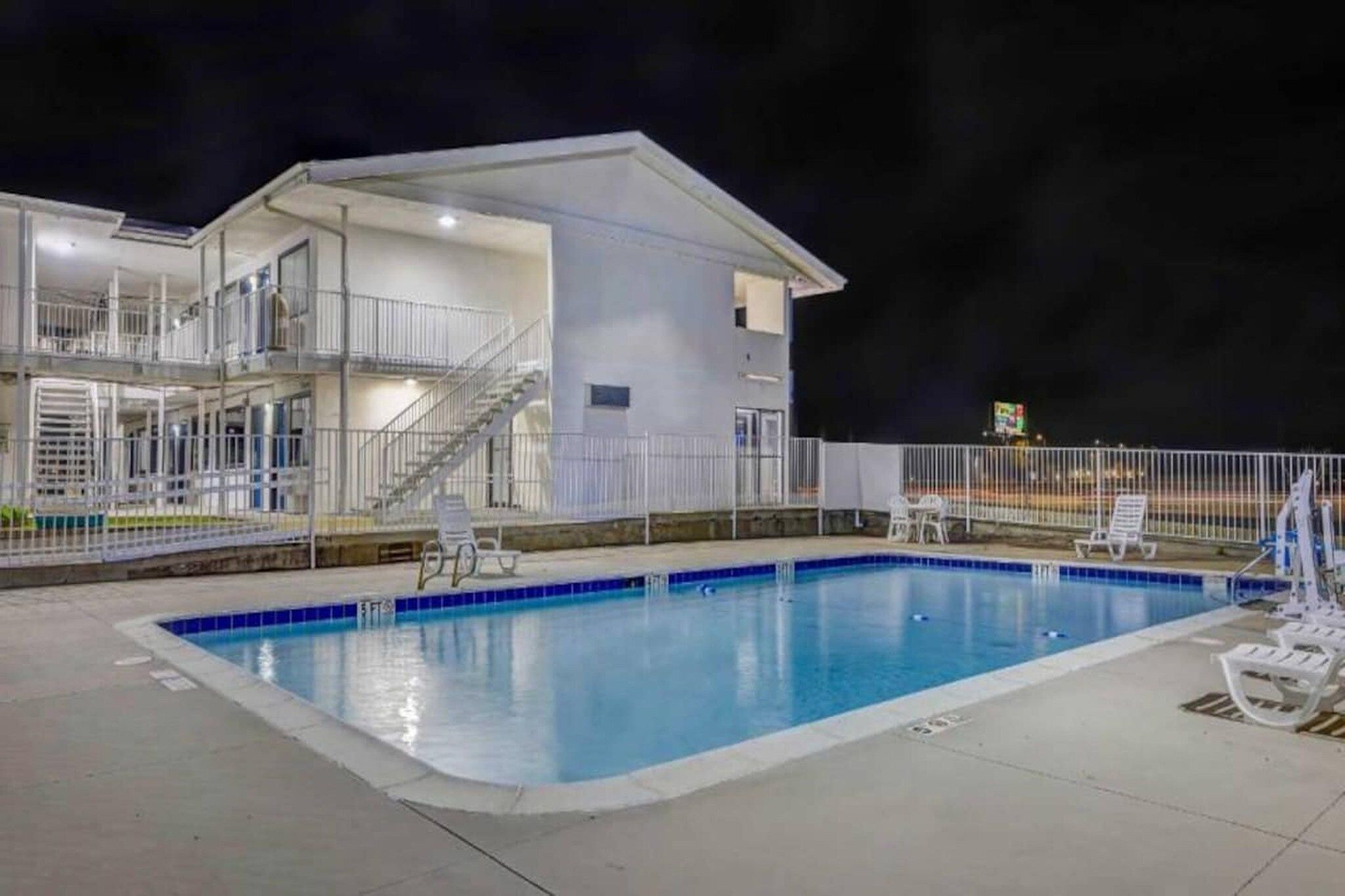 A swimming pool with a white building adjacent to it, featuring a staircase and a fenced area with lounge chairs.
