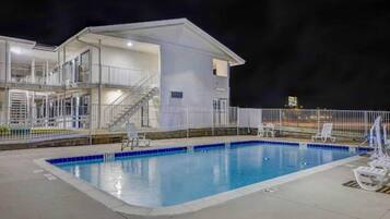 A swimming pool with a white building adjacent to it, featuring a staircase and a fenced area with lounge chairs.